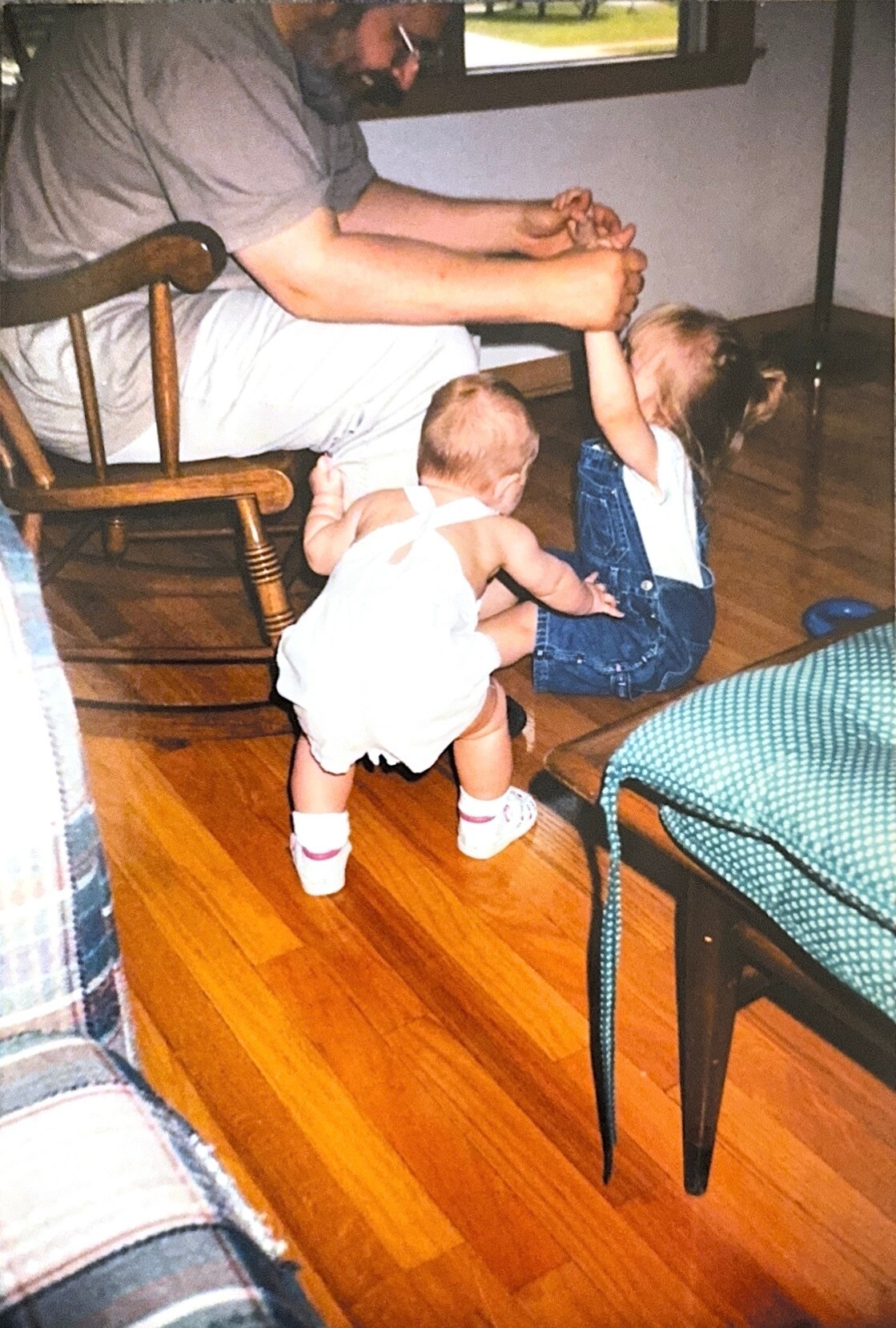 Man with a beard sits on a chair, holding the hands of a girl in denim overalls as a crawling toddler reaches nearby on a wooden floor.