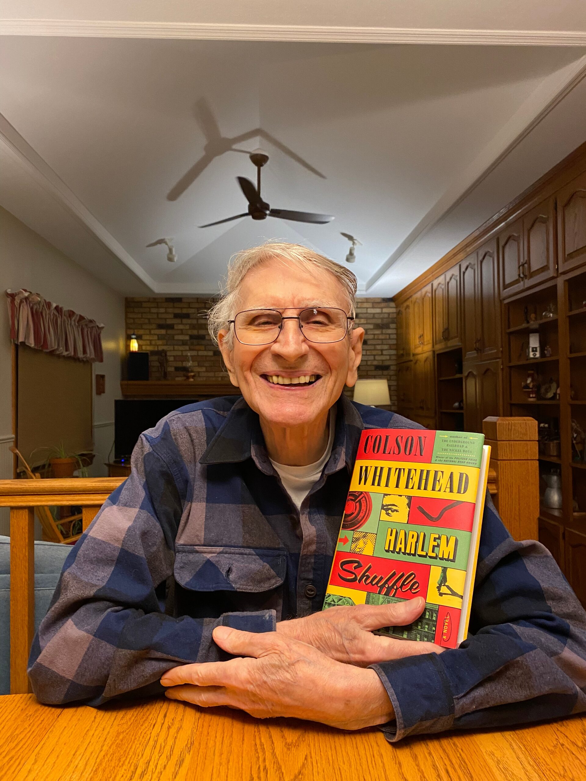 Smiling elderly man with glasses sitting at a wooden table, holding a colorful book titled Harlem Shuffle by Colson Whitehead in a home kitchen.