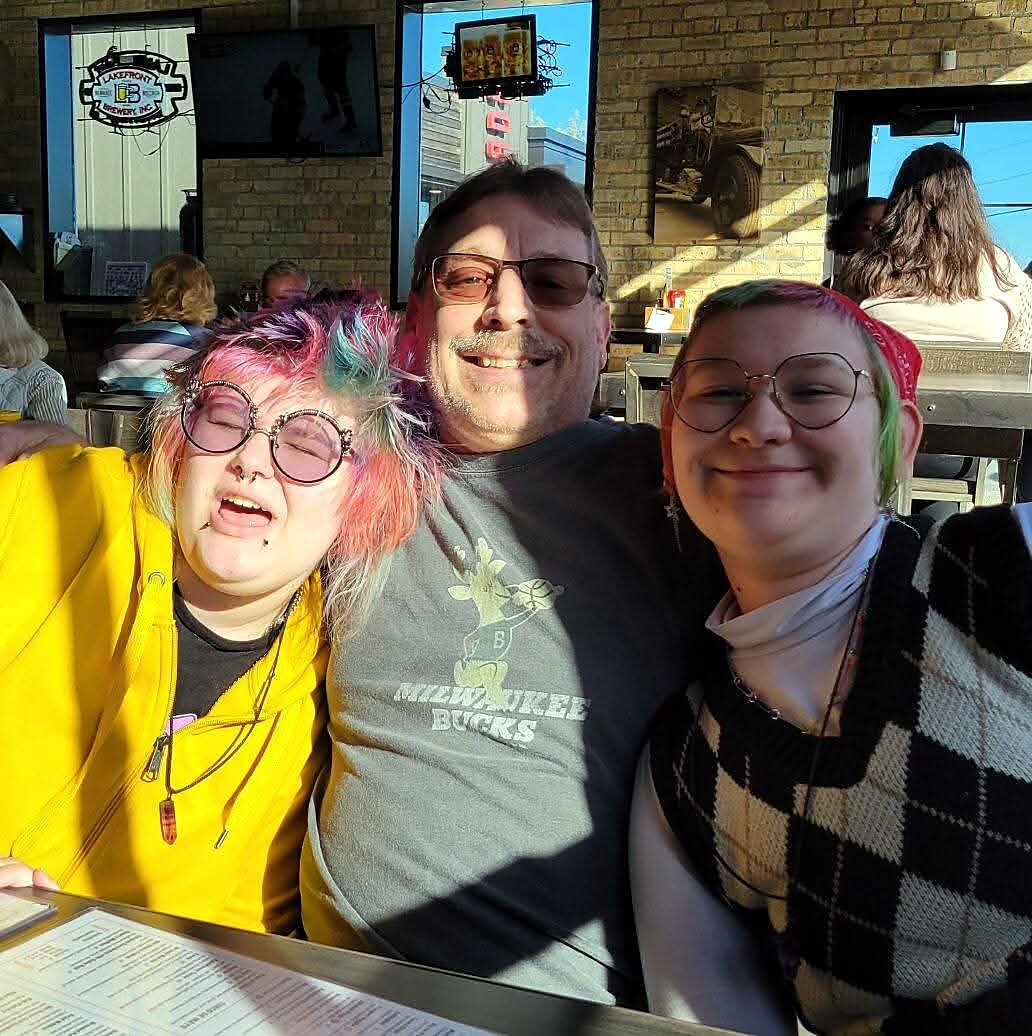 Three friends pose for a photo at a sunlit restaurant, left person with colorful hair and yellow jacket, center man in a gray shirt, right person with colorful hair and a black‑and‑white checkered sweater.