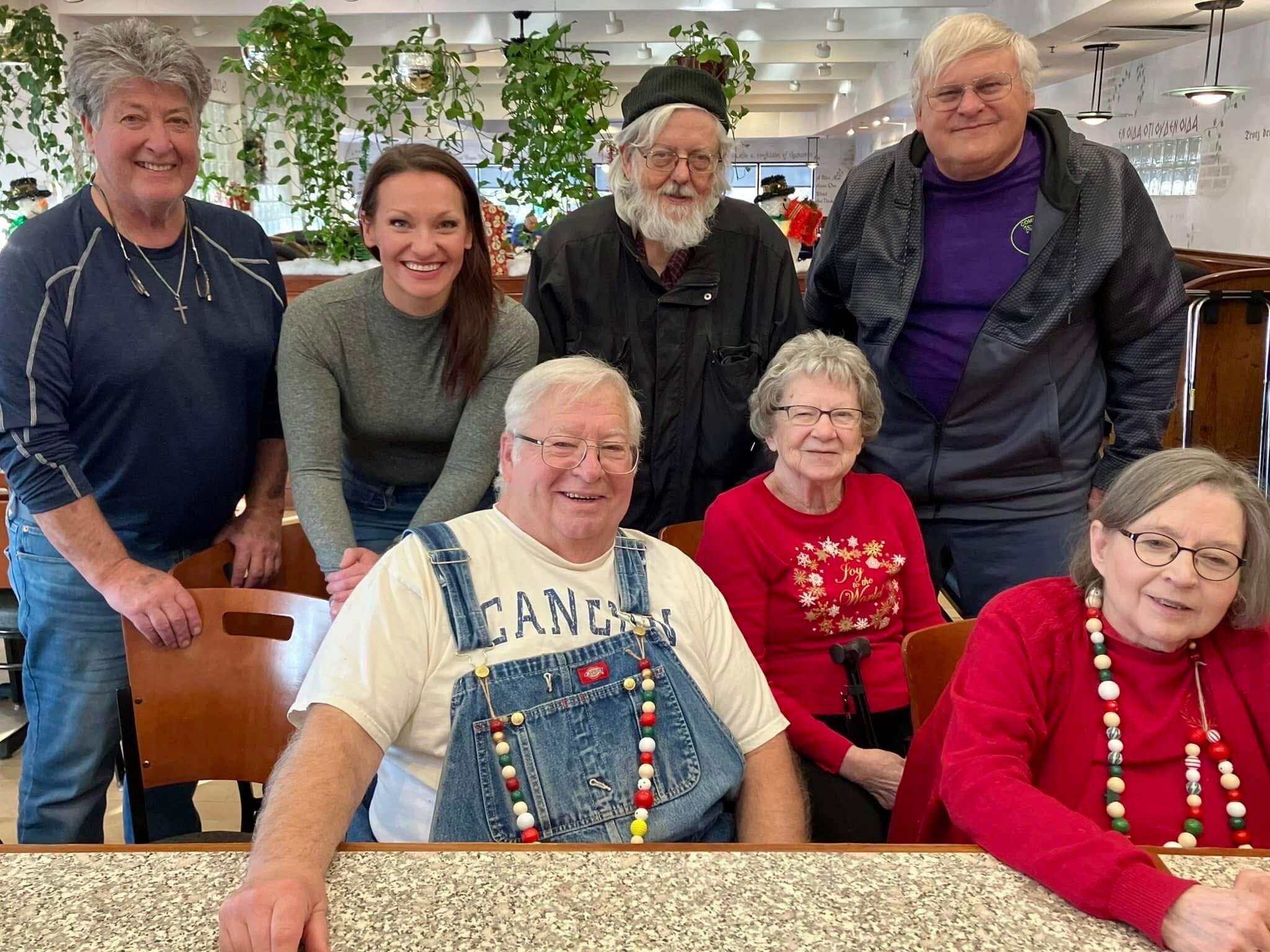 Group of seniors posing for a photo at a festive gathering in a bright dining area, smiling and wearing colorful bead necklaces.