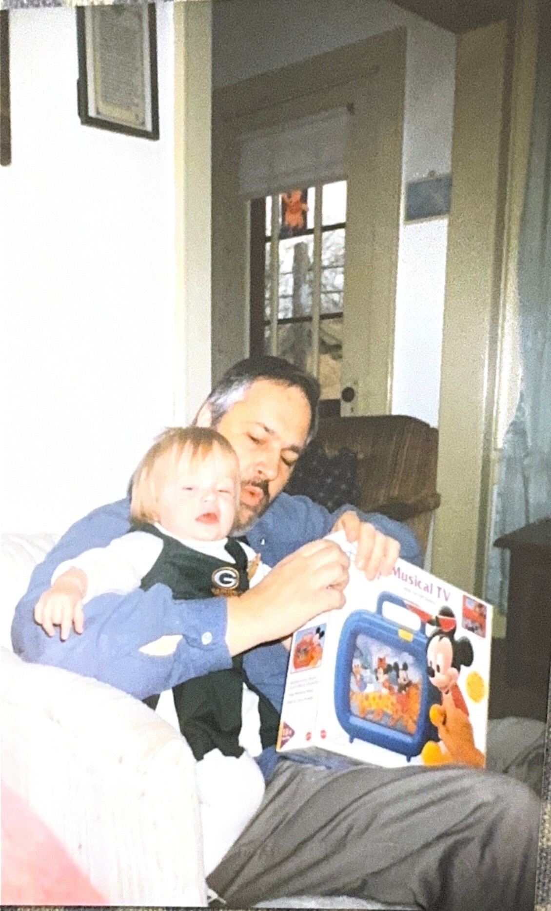 Father and toddler sitting on a sofa, playing with a Mickey Mouse toy box or TV-themed toy game.