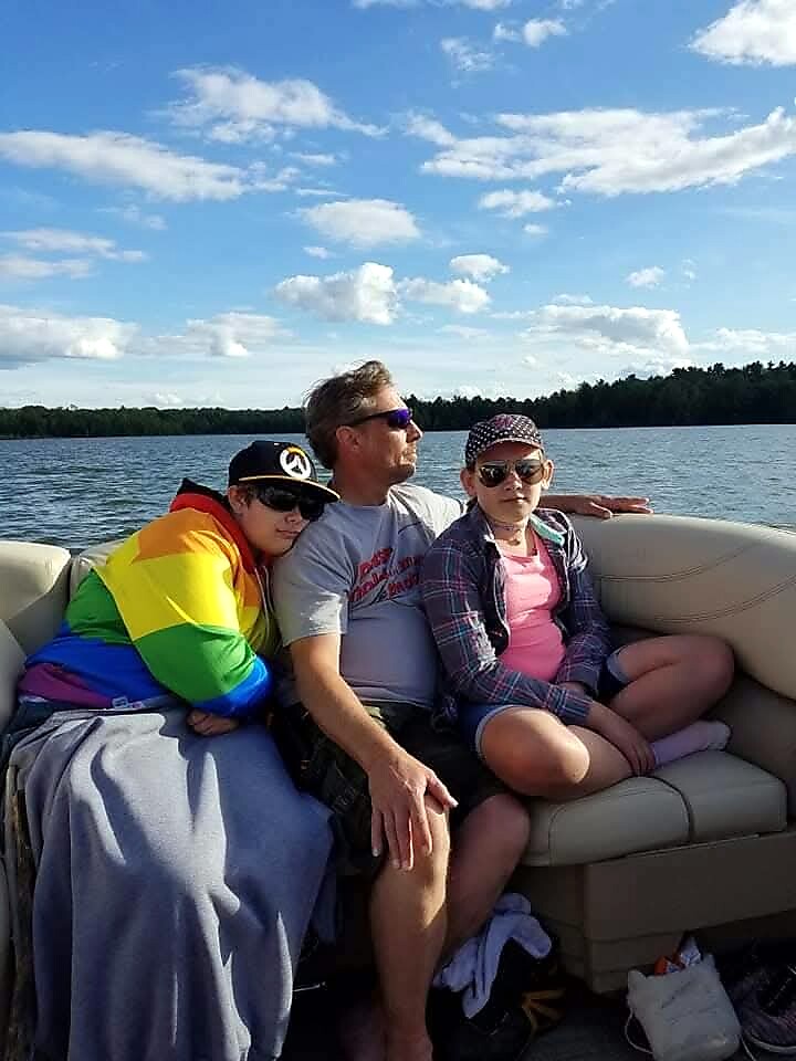 A man and two children sit on a beige boat seat on a sunny lake, smiling at the camera.