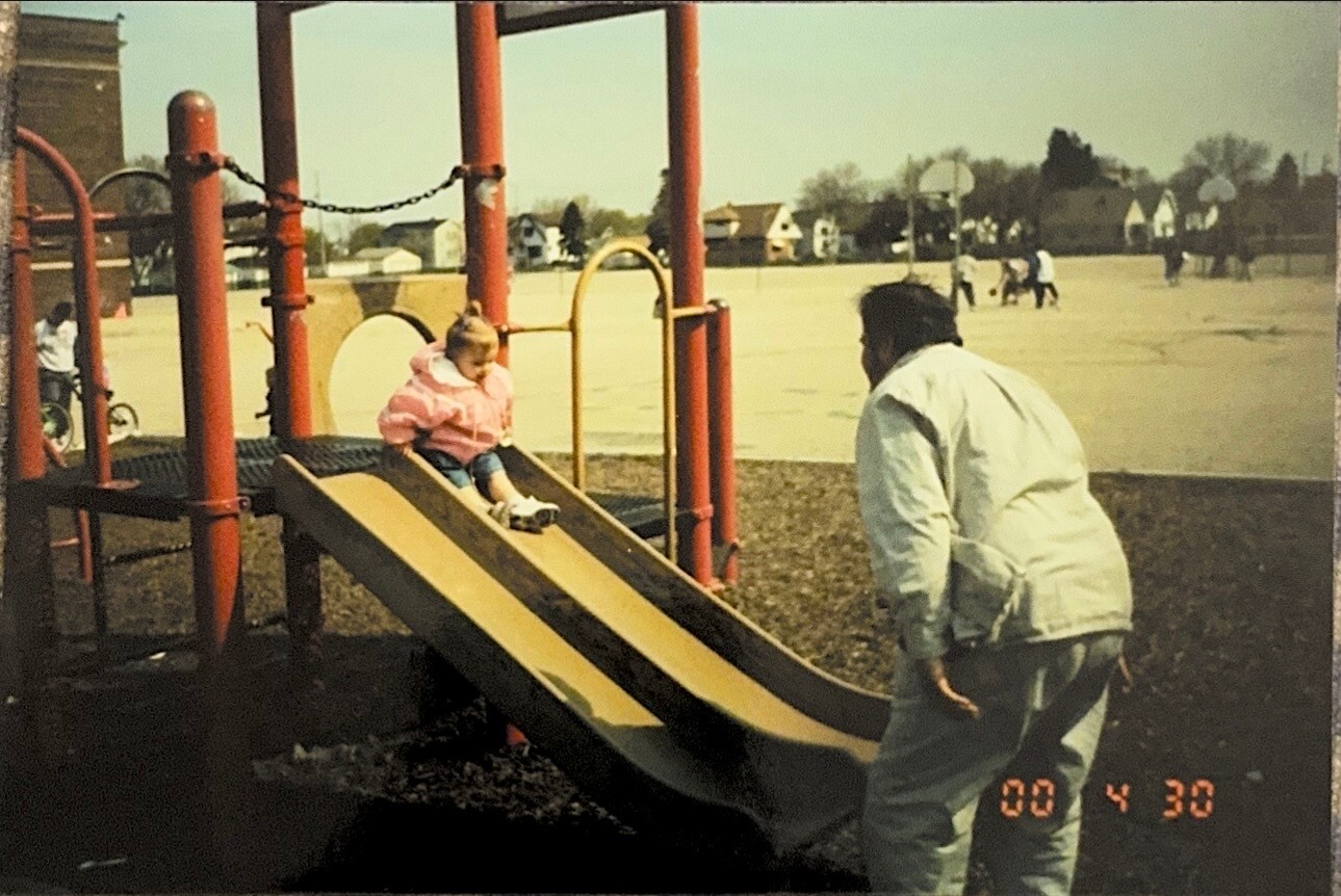 Child in a pink jacket sliding down a beige playground slide while an adult in a light jacket approaches from the right in a park setting.