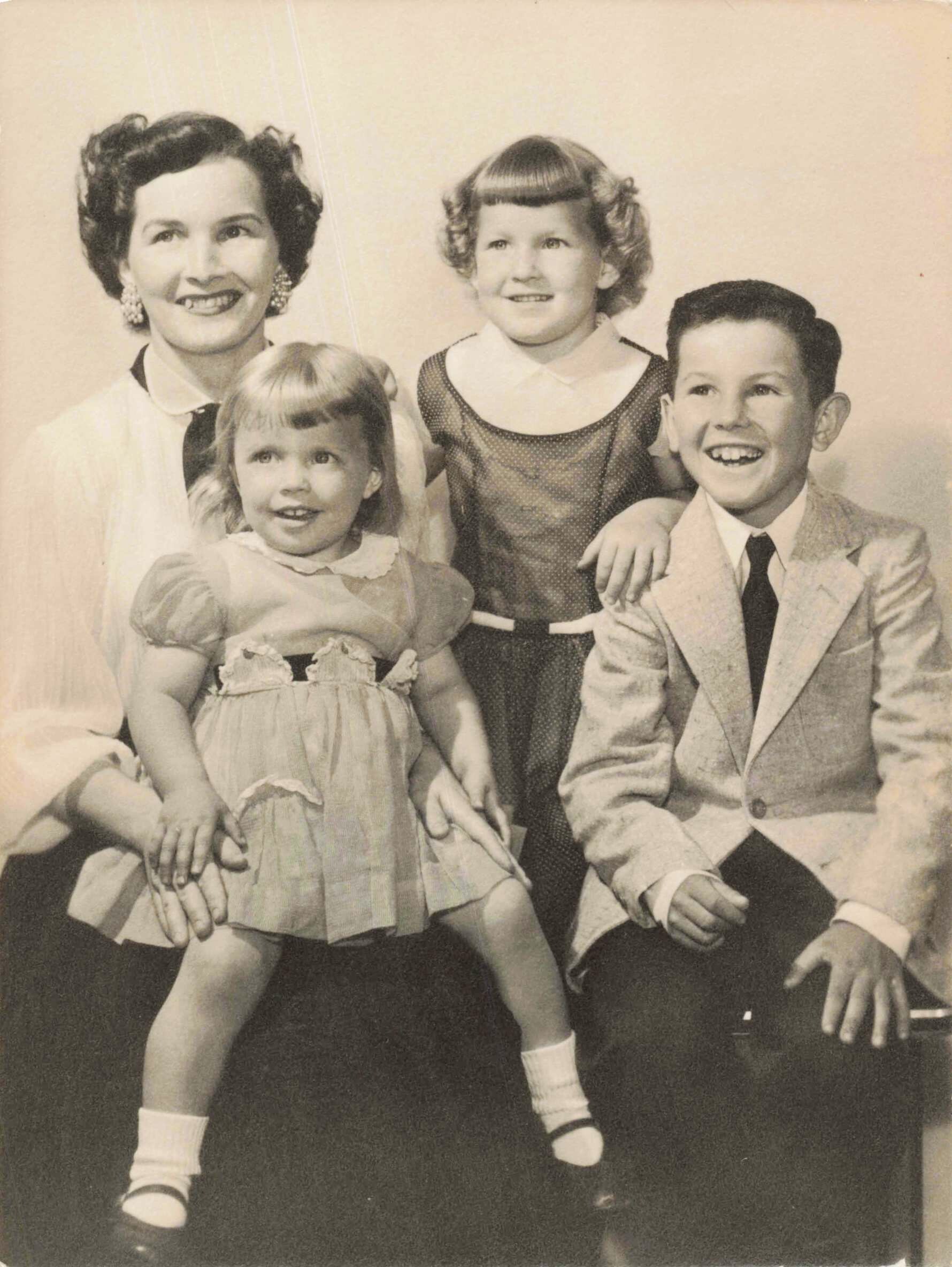 Mom poses with her three kids: two girls and a boy, all smiling in a vintage studio portrait.