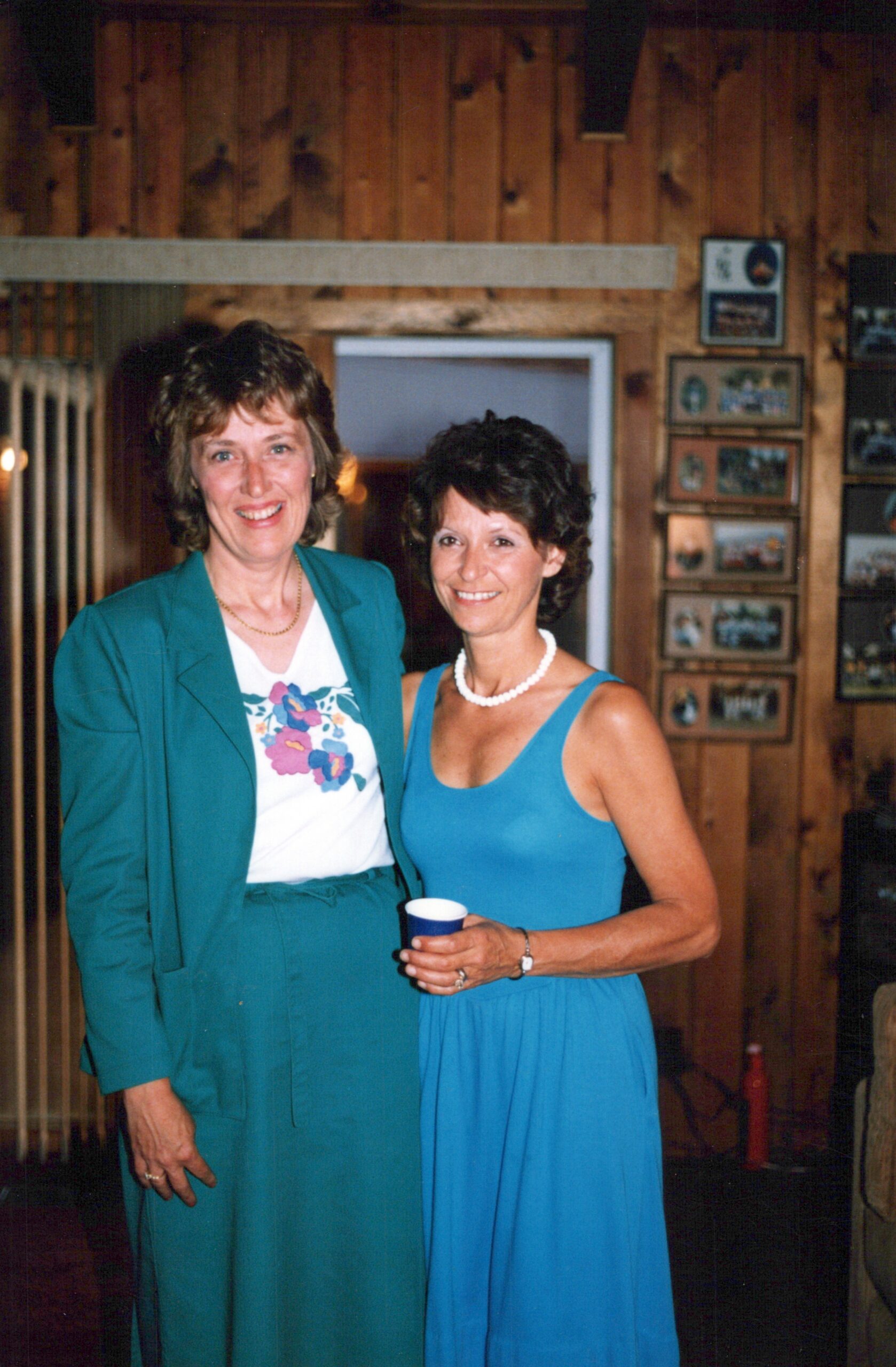 Two women smile for a photo indoors, one in teal suit and floral shirt, the other in a blue dress, holding a cup, with wooden wall and memorabilia in the background.
