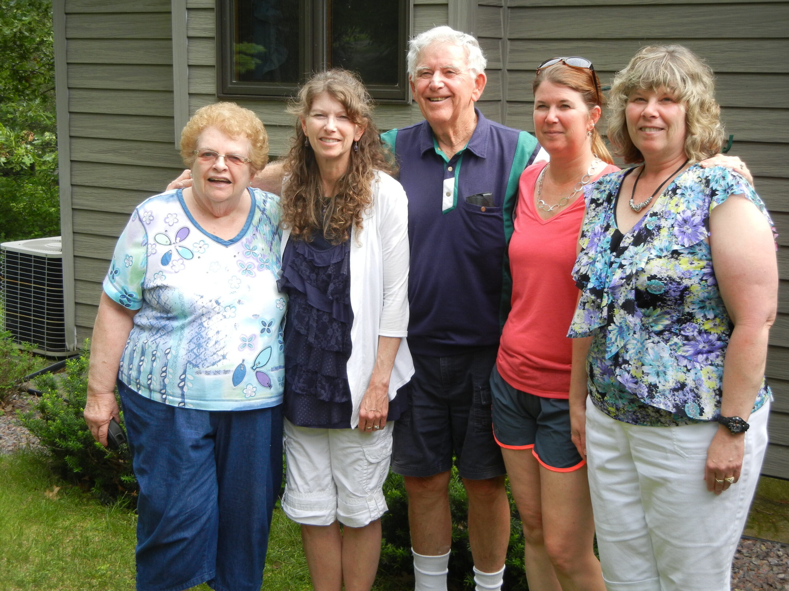 Five adults posing together outside a wooden house, smiling at the camera.