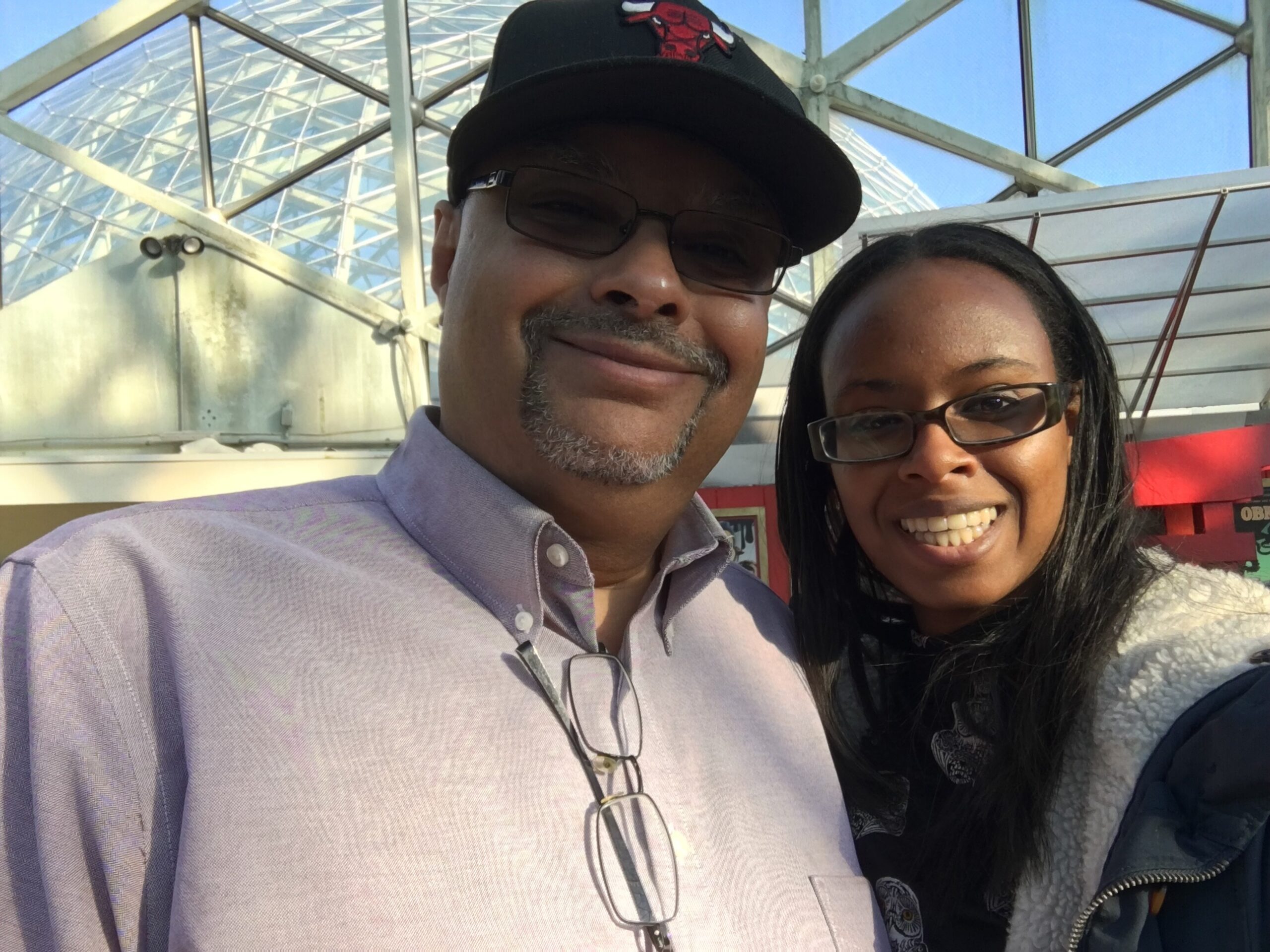 Smiling man wearing a black Chicago Bulls cap and glasses, posing with a woman wearing glasses outdoors against a glass-and-steel structure.