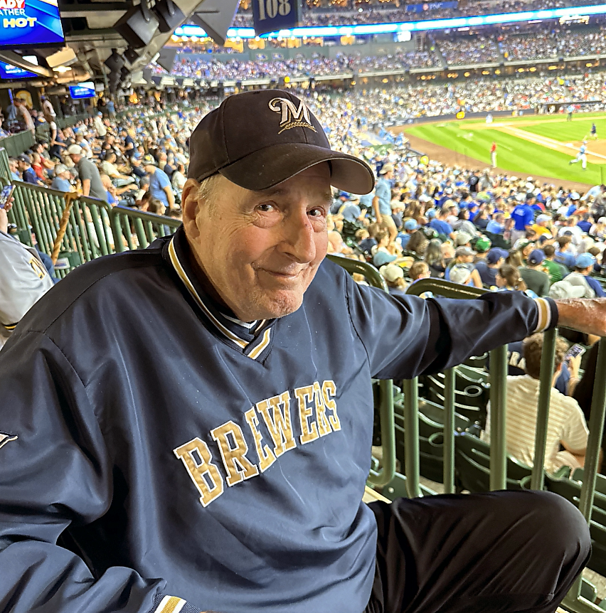Older man in a Brewers cap and navy Brewers jersey sits in stadium seating, smiling with the baseball field and crowd in the background.