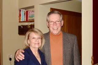 Smiling older couple standing indoors; woman in a blue blazer and the man in a brown turtleneck with a gray blazer.
