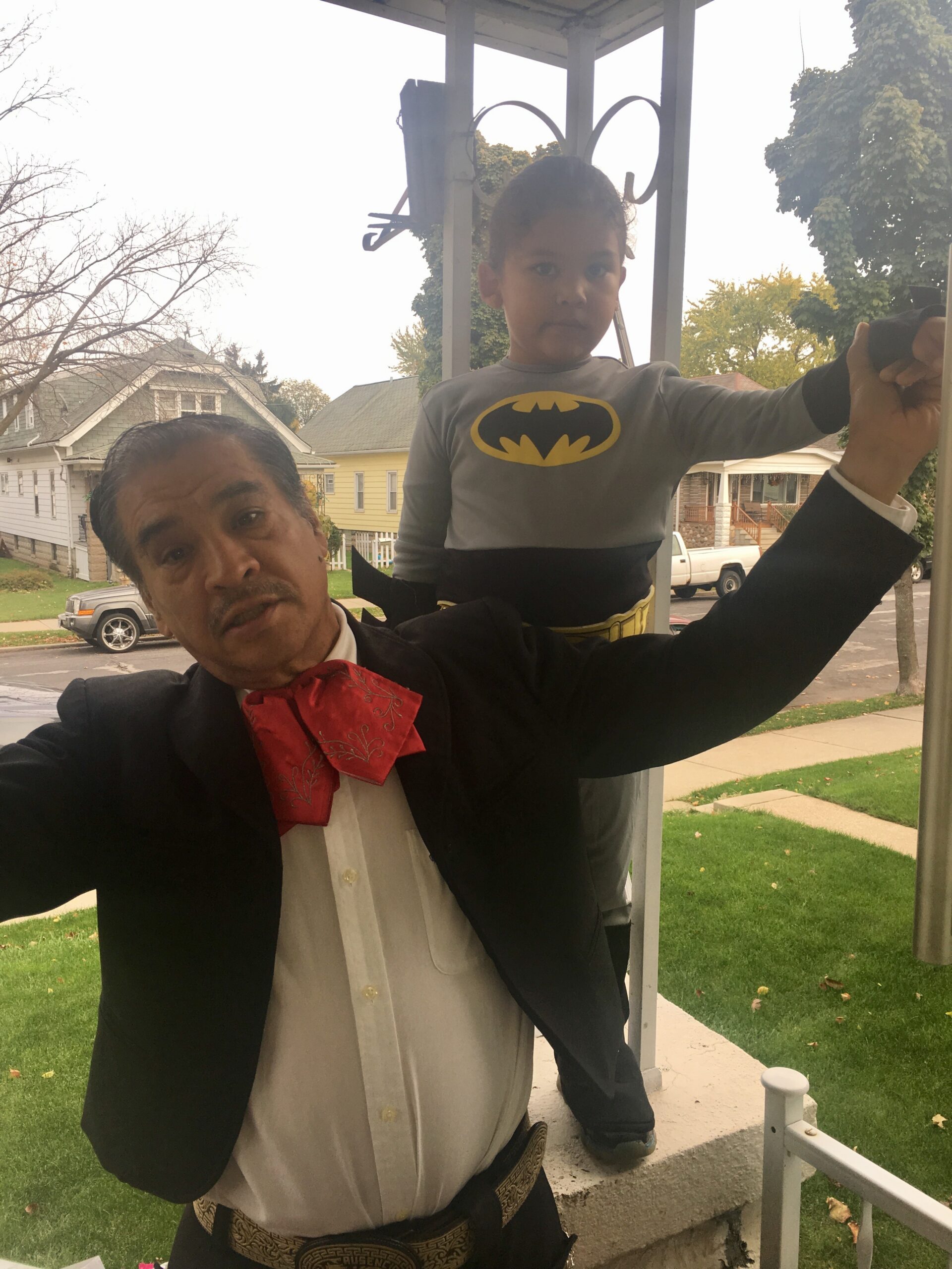 Adult man in a suit and red bow tie lifts a child onto his shoulder on a residential porch outdoors; child wears a Batman shirt.