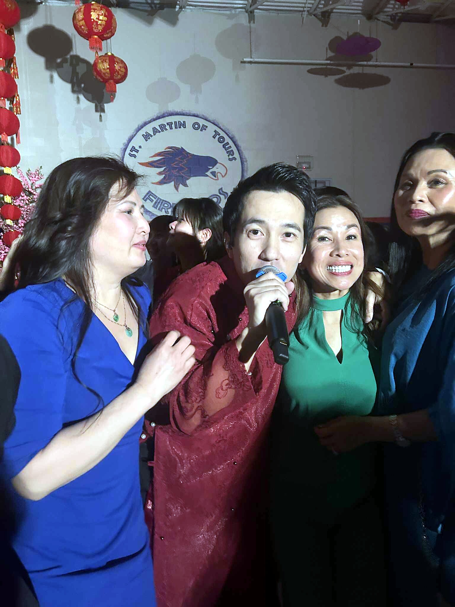Group of people at a festive event singing into a microphone, with red lanterns and a circular logo in the background.