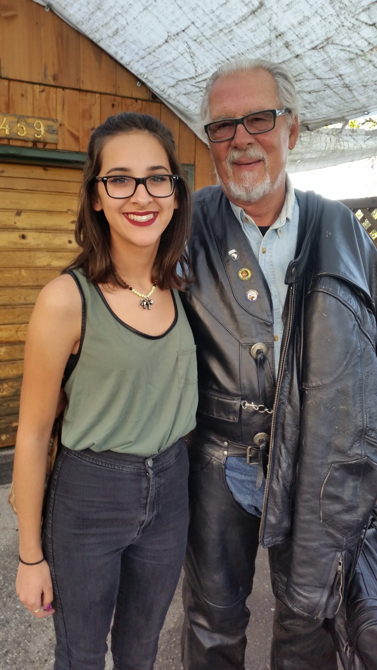 Two smiling people posing together outdoors: a young woman with glasses beside an older man in a leather jacket in front of a wooden building.