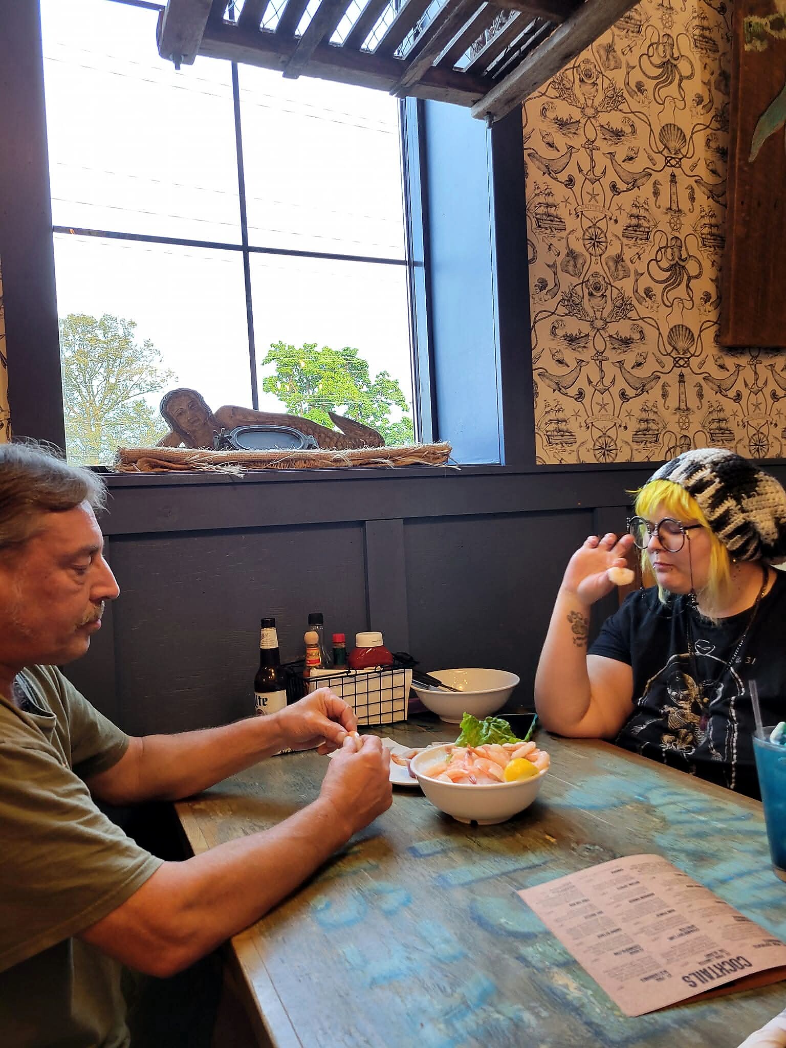 Two people sit at a weathered wooden table, eating salmon sashimi bowls with sauces and drinks in front of them near a large window.