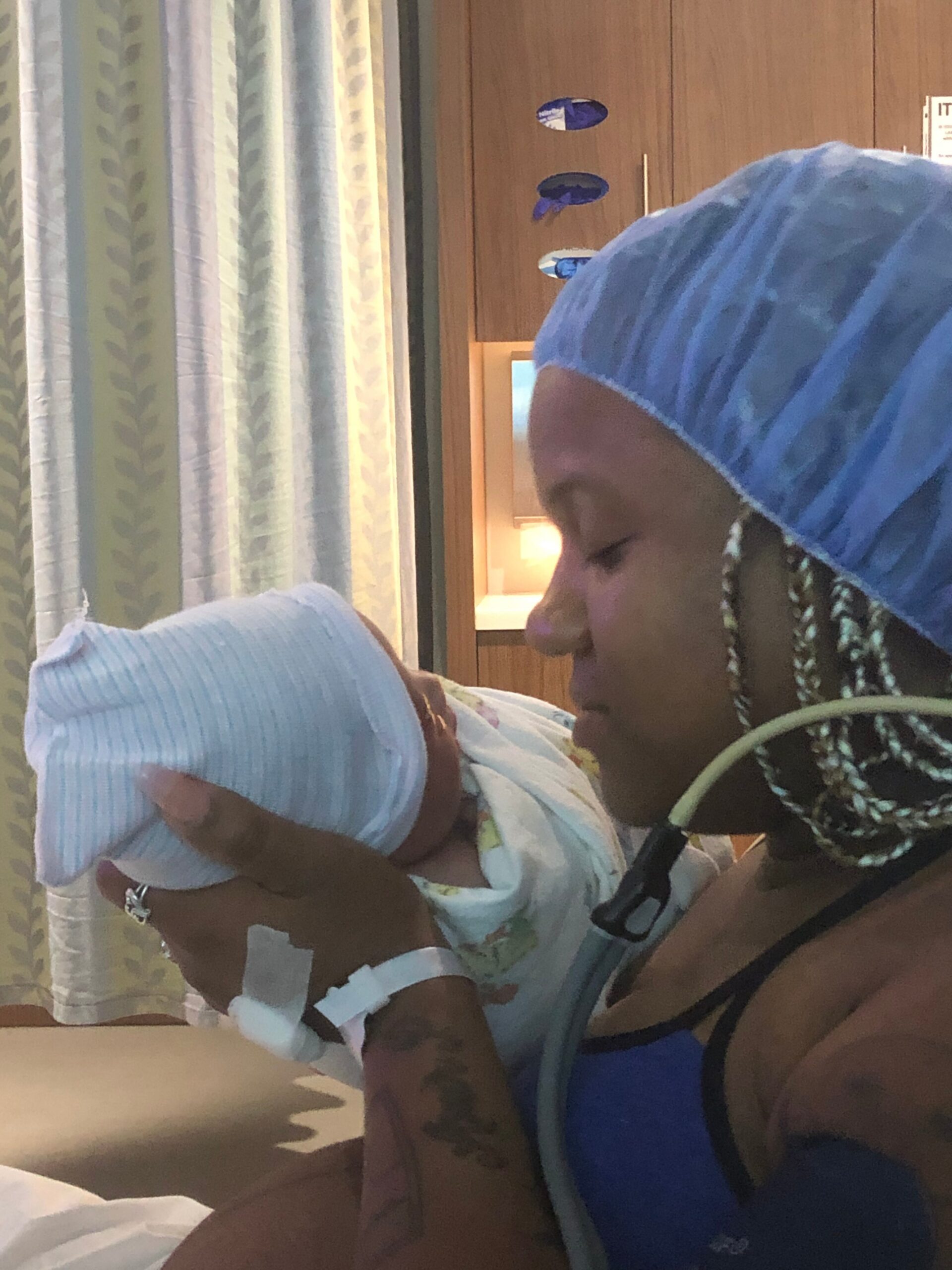 Nurse wearing a blue surgical cap cradles a newborn wrapped in a blanket as a parent in a blue striped cap leans in, in a hospital room.