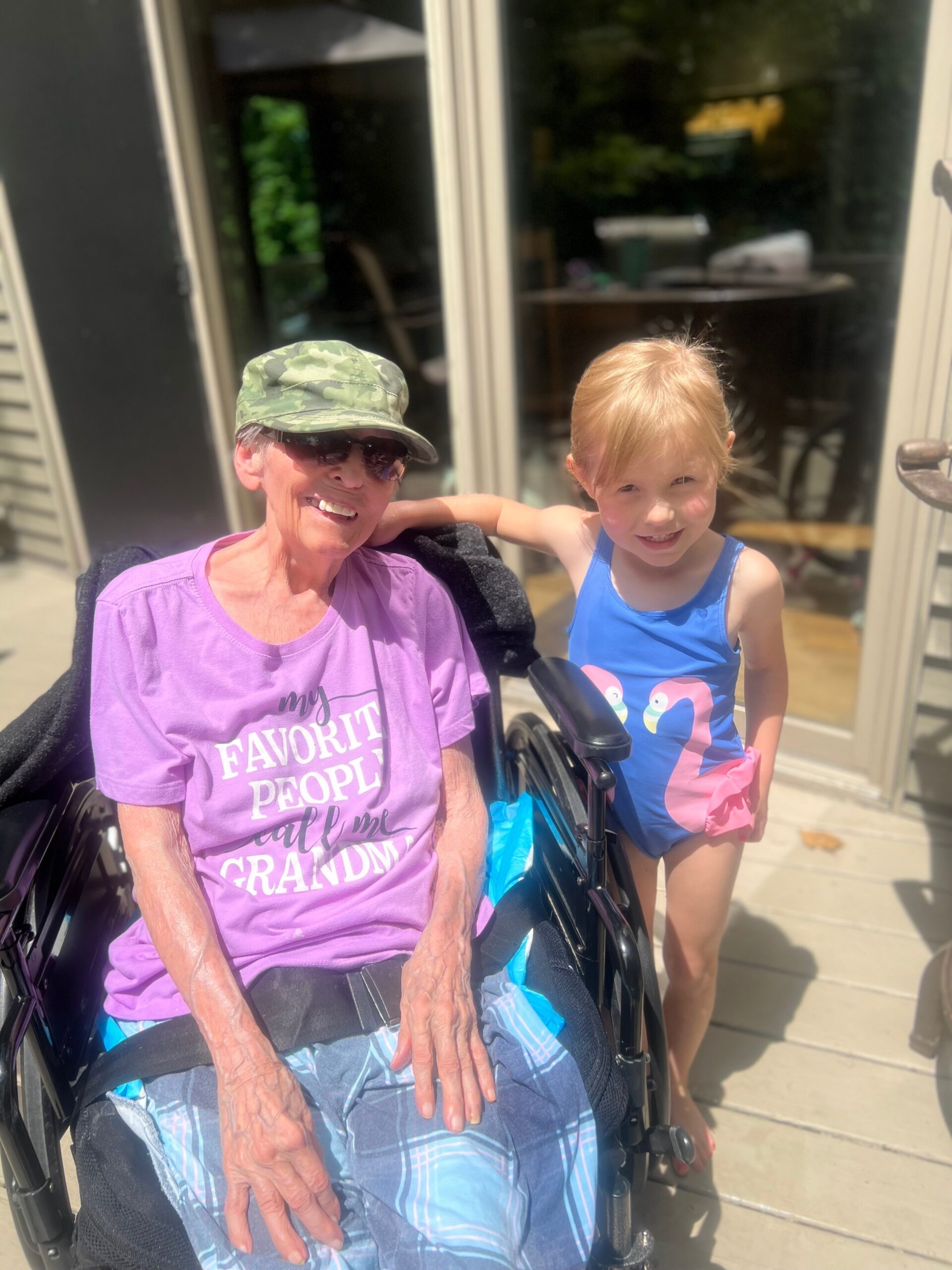 Grandmother in a wheelchair smiles beside a girl in a blue swimsuit on a sunny porch.