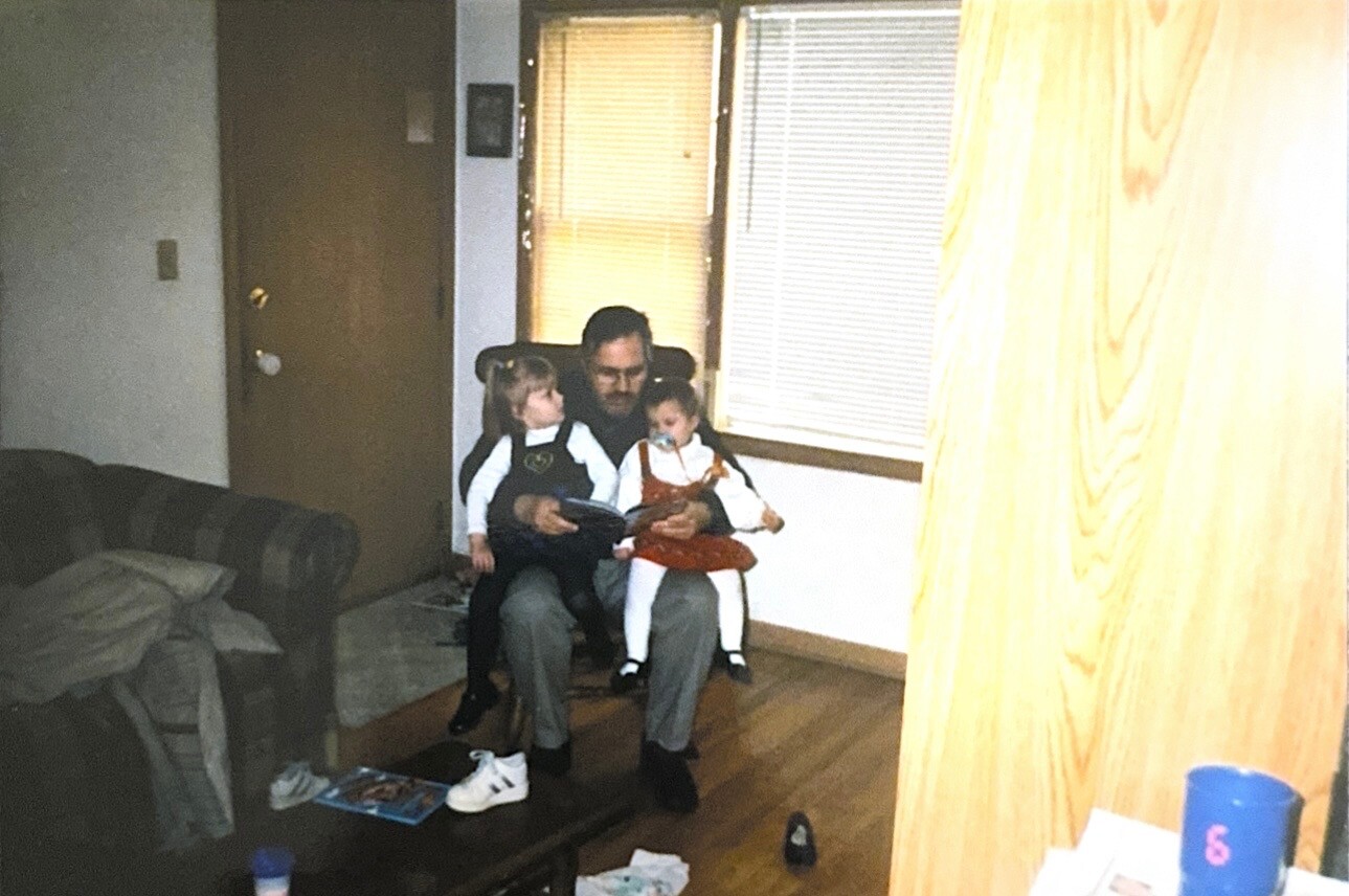 A man sits in a living room with two young girls on his lap, reading a book together.