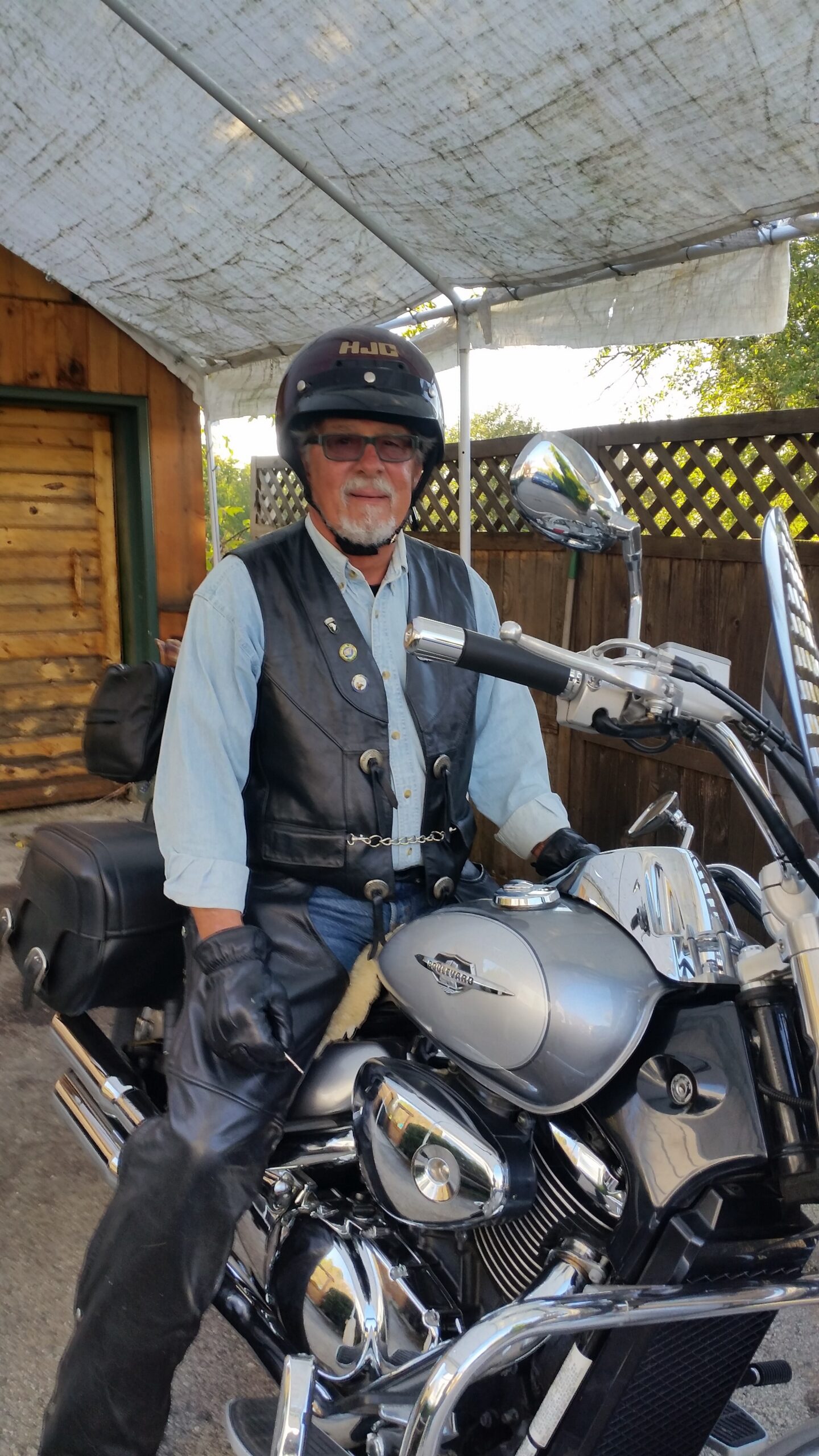 Older man with a white beard wearing a black helmet and leather vest sits on a silver motorcycle in a driveway under a canopy.