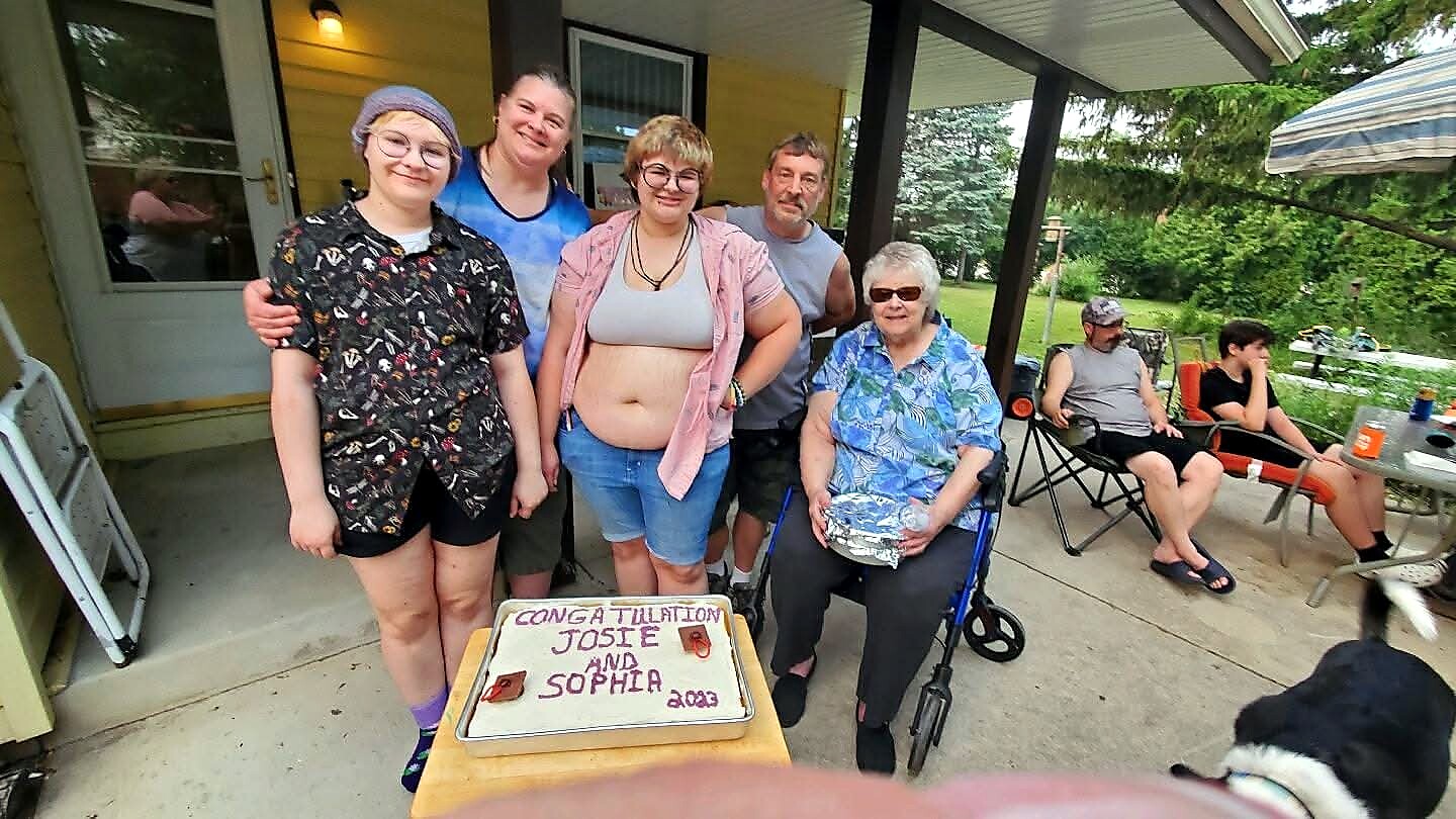 Group of people posing on a porch for a celebratory photo with a cake on a table in front; others sit nearby under a canopy and trees in a yard option.