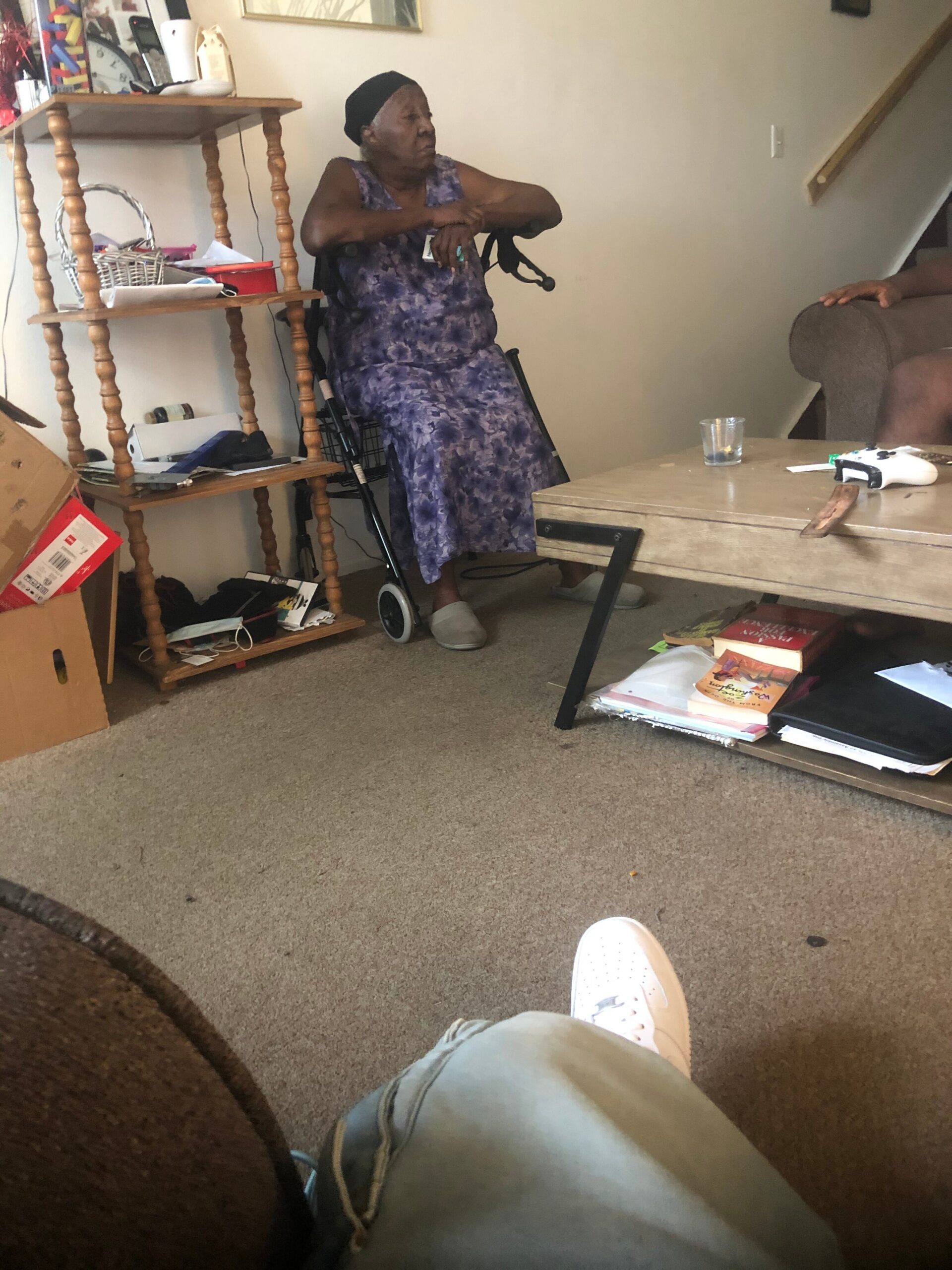 Older woman in a purple patterned dress sits in a wheelchair in a cluttered living room, with a walker nearby and a wooden coffee table in front.