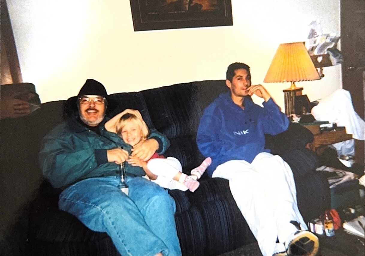 Three people sit on a dark couch in a living room: an older man in a beanie with a beer, a smiling child on his lap, and a teen in a blue hoodie nearby.