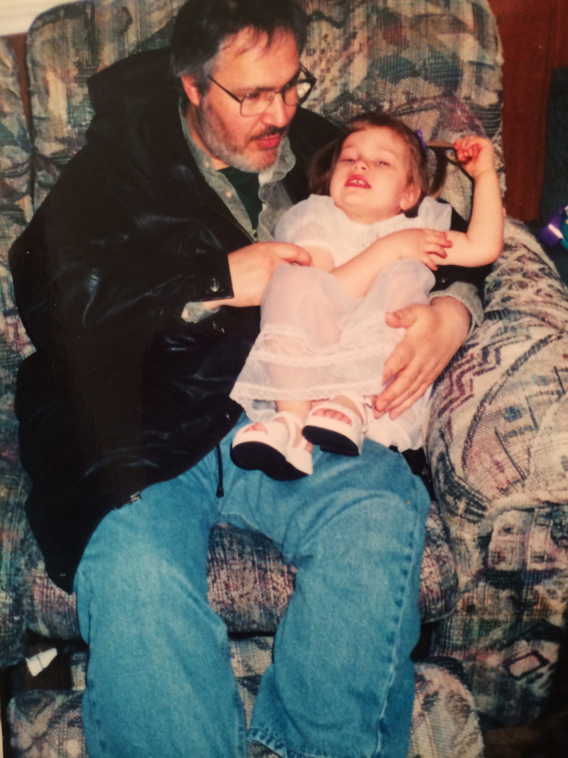 Man with glasses and a grey beard sits on a patterned sofa, holding a young girl in a white dress on his lap while he adjusts her hair.