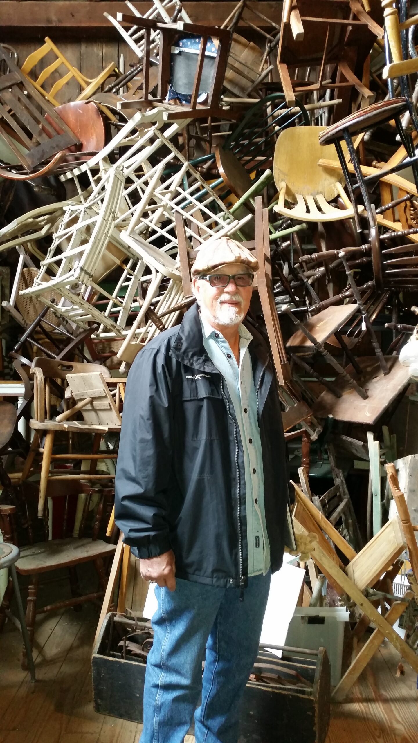 Older man wearing a cap and glasses stands in front of a chaotic pile of wooden chairs in a workshop.