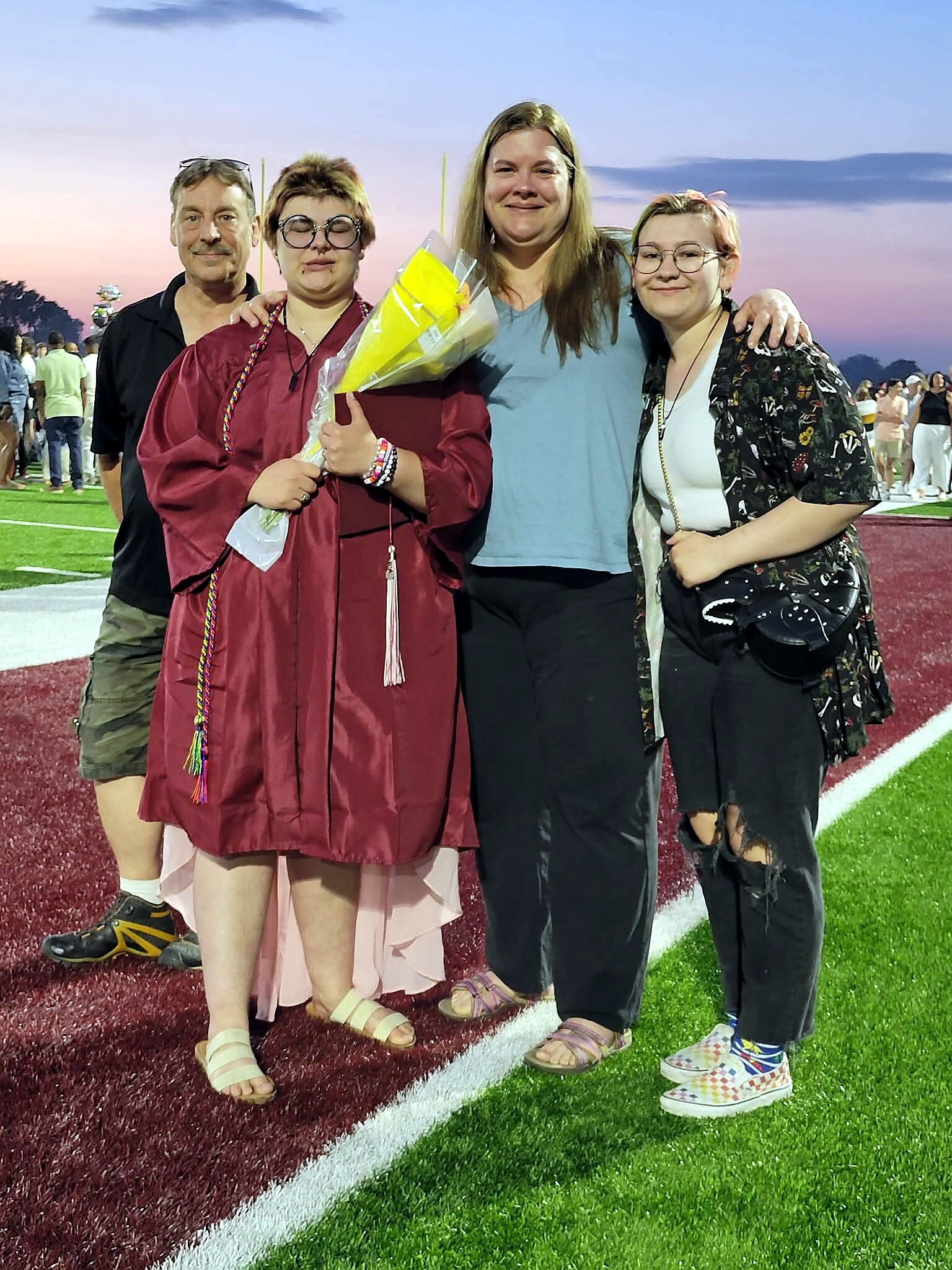 Four people pose on a track at sunset; a graduate in a maroon gown holds a bouquet, flanked by family and friends.