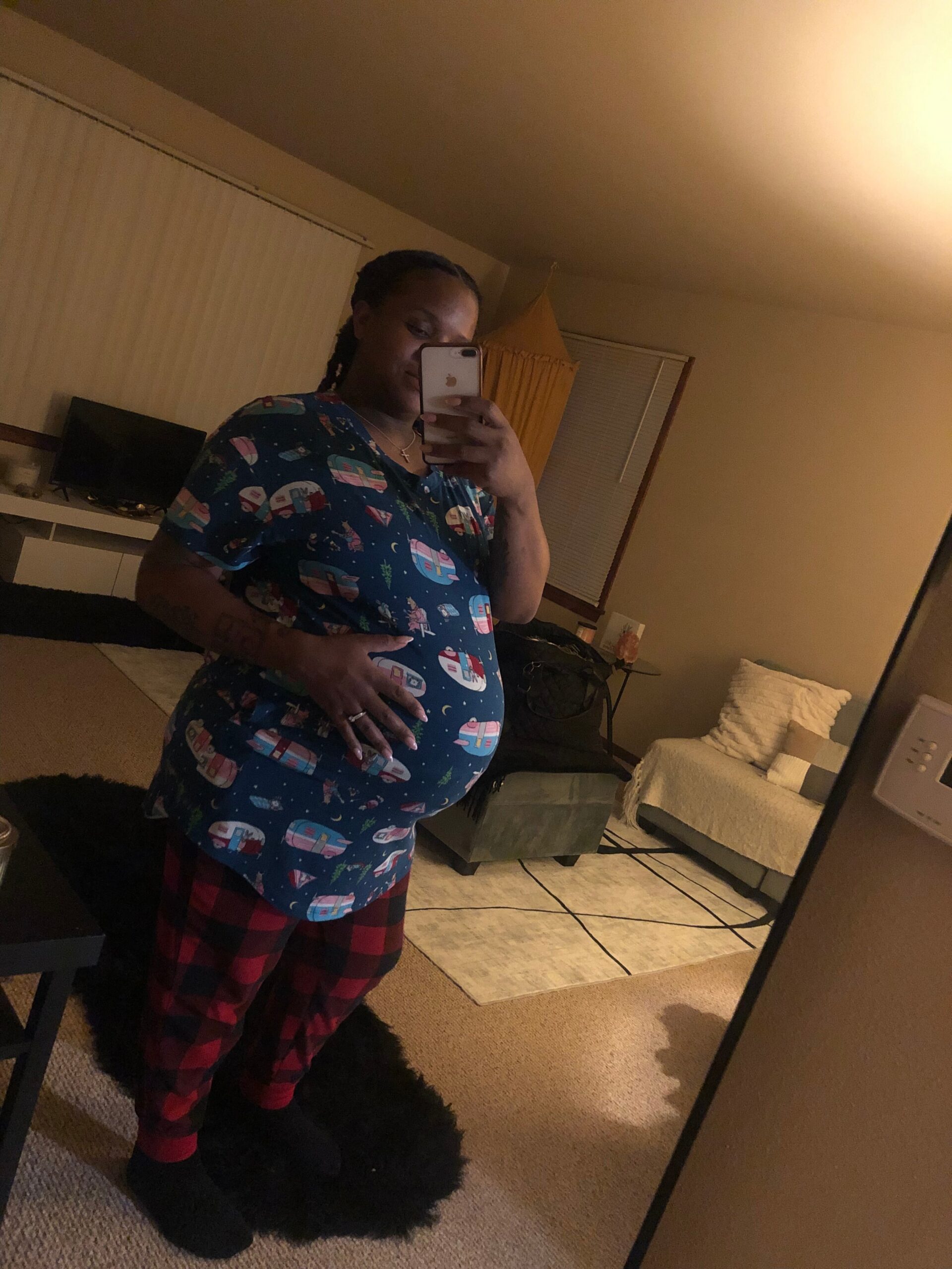 Pregnant person taking a mirror selfie in a bedroom, wearing a blue space-themed top and red plaid pants, smiling at the camera.