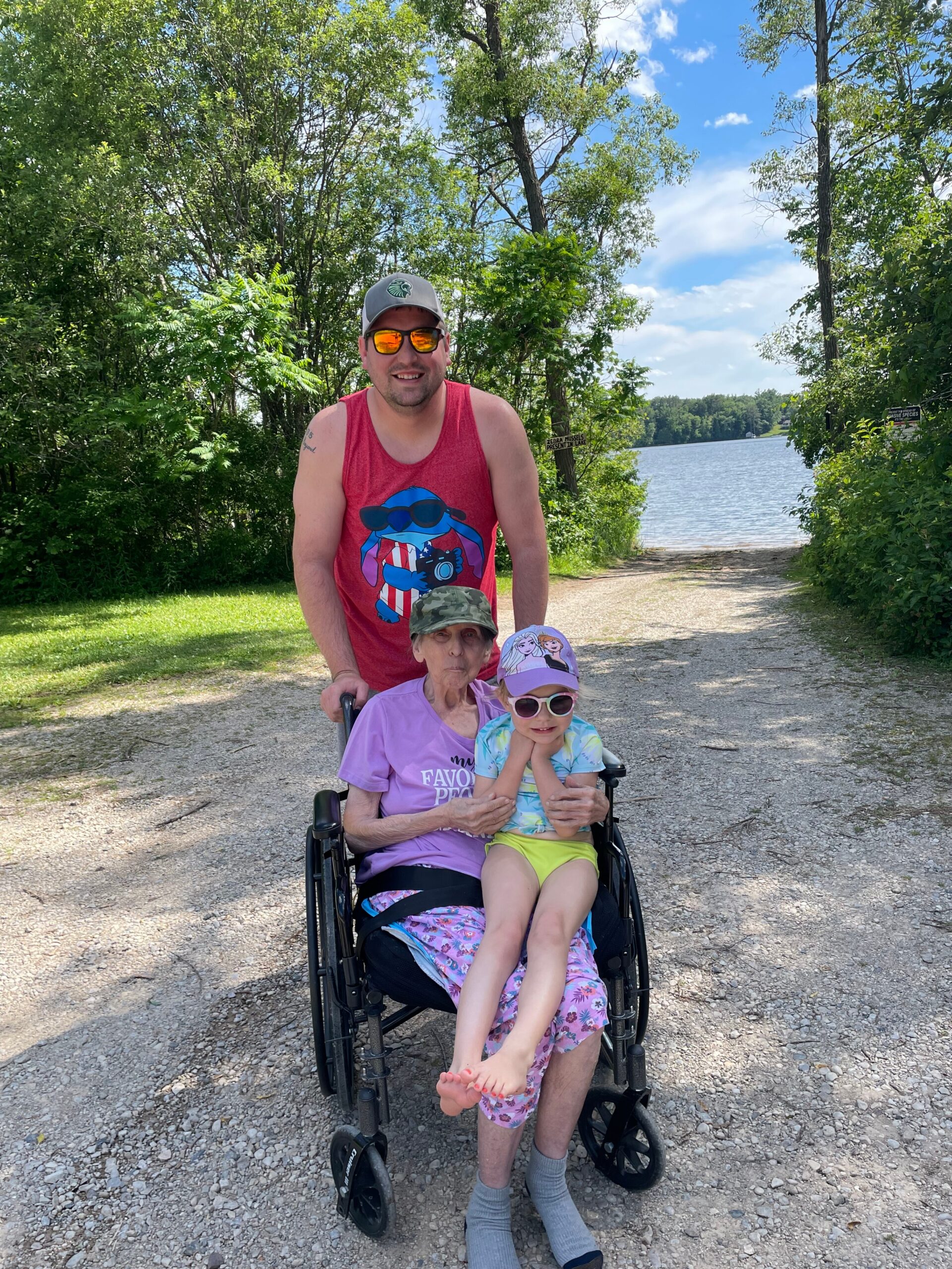 Man pushing a wheelchair with an elderly woman and a young girl along a lakeside path on a sunny day.