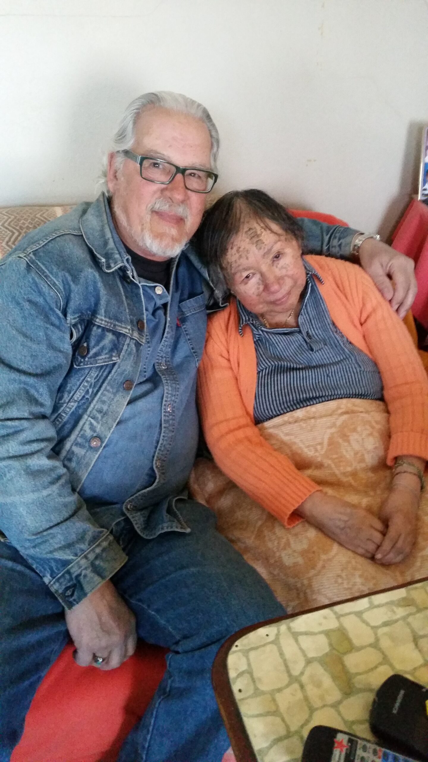 Smiling elderly man in glasses and denim jacket sits with his arm around an elderly woman in an orange cardigan on a patterned sofa at home.