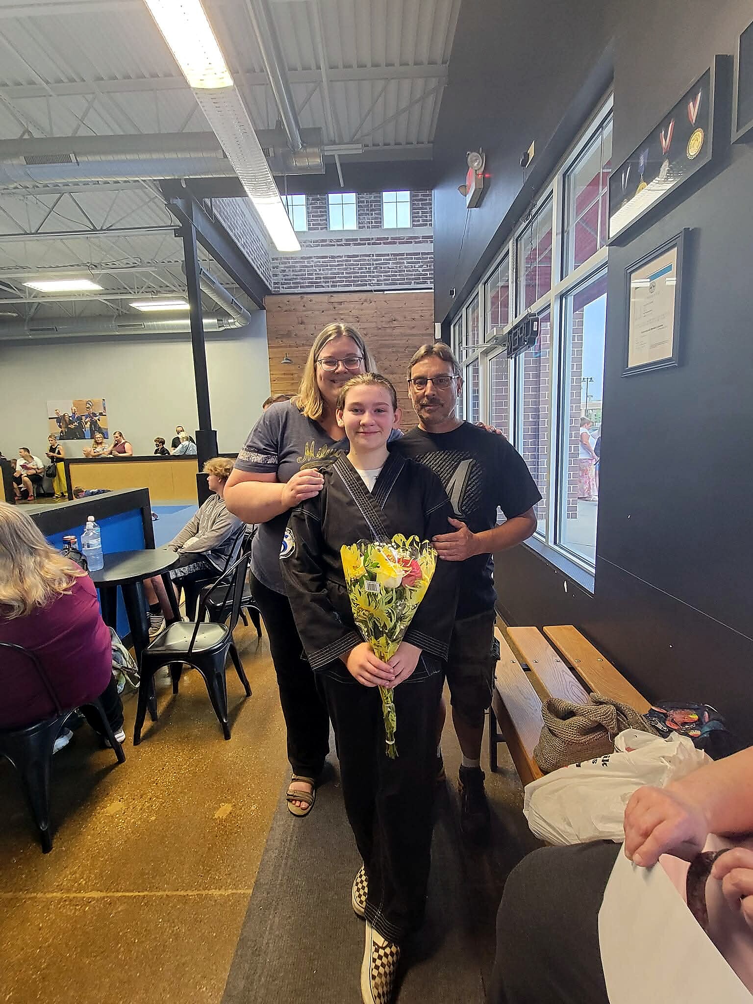 Girl in a black martial arts gi holds a bouquet, flanked by two adults in a gym or dojo setting with others in the background.