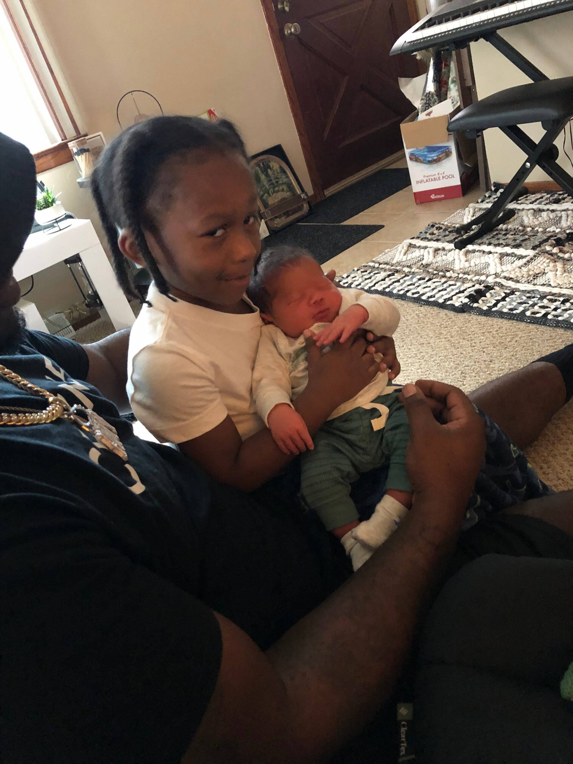 Adult man with braided hair cradling a sleeping newborn baby in a living room interior.