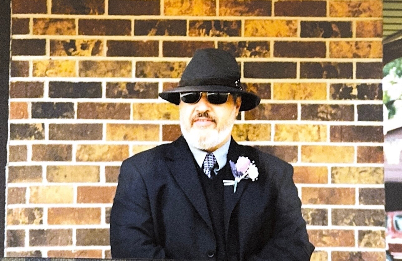 Man in a black suit, fedora, and sunglasses standing against a brick wall with a pink boutonniere.