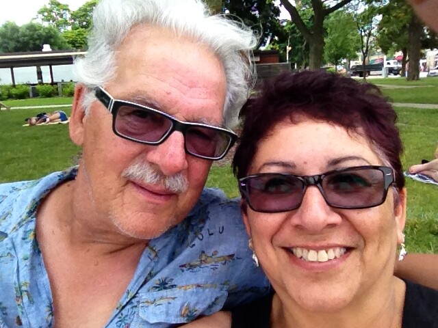 Smiling older couple wearing sunglasses pose for a selfie in a sunny park; man with white hair and mustache in a tropical shirt, woman with burgundy hair in a black top.