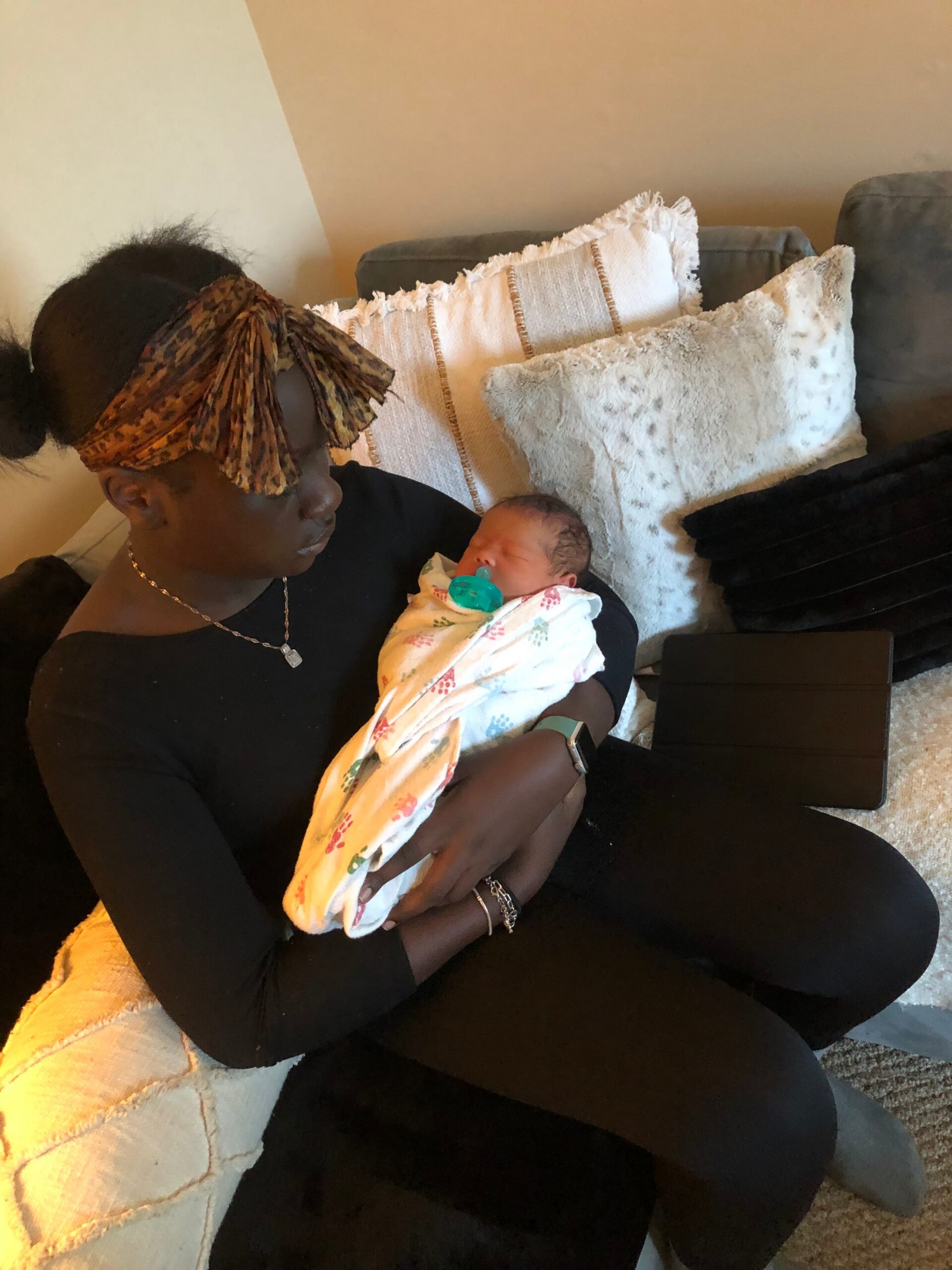 Woman wearing a leopard print head wrap cradling a sleeping newborn swaddled in a floral blanket on a beige couch.