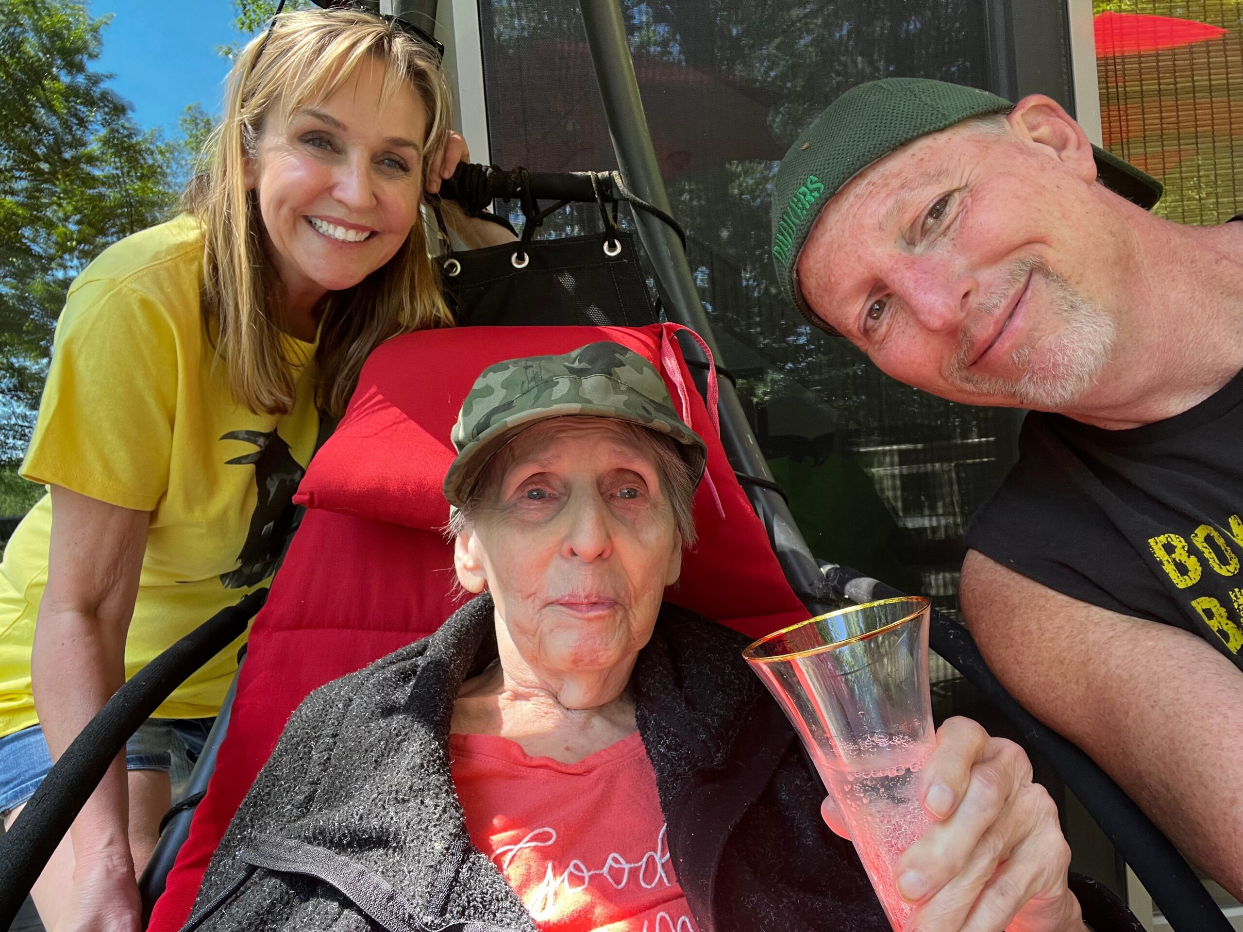 Three people posing for a sunny outdoor selfie: a woman in a yellow shirt on the left, an elderly woman in a red jacket and camo hat in the center, and a man in a green cap leaning in with a pink cocktail in the foreground.