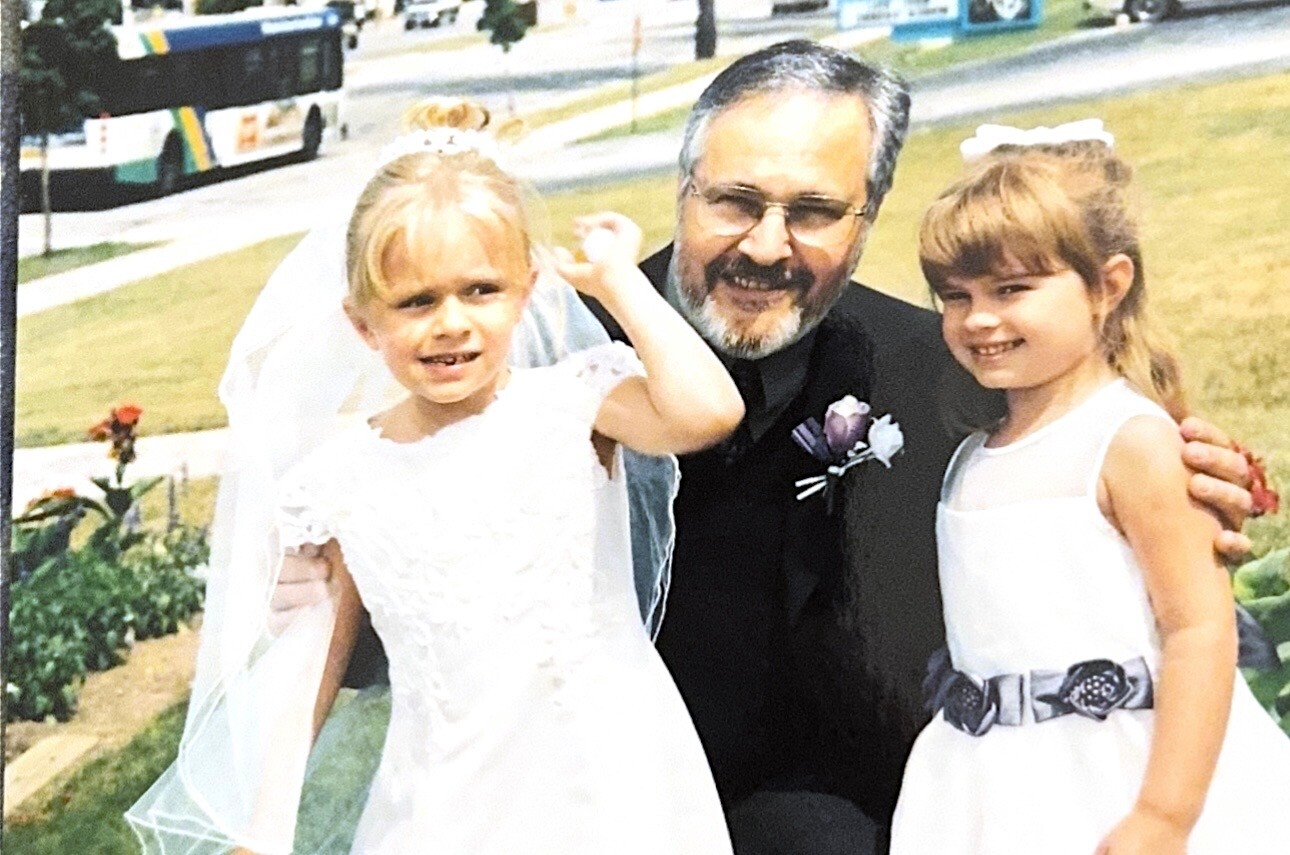 Two young girls in white dresses pose with a smiling man in a suit outdoors at a wedding. One girl wears a veil and the other a silver belt.