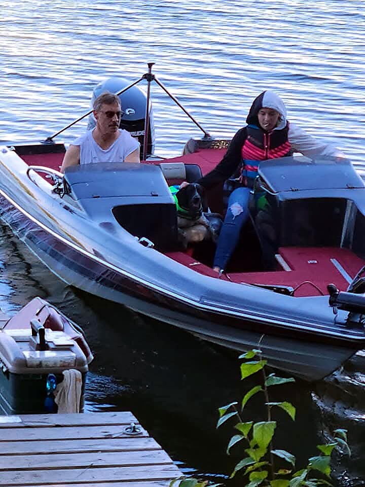 Two people sit in a red and gray speedboat docked at a wooden pier by calm water, one wearing sunglasses and the other in a hoodie with a life jacket.