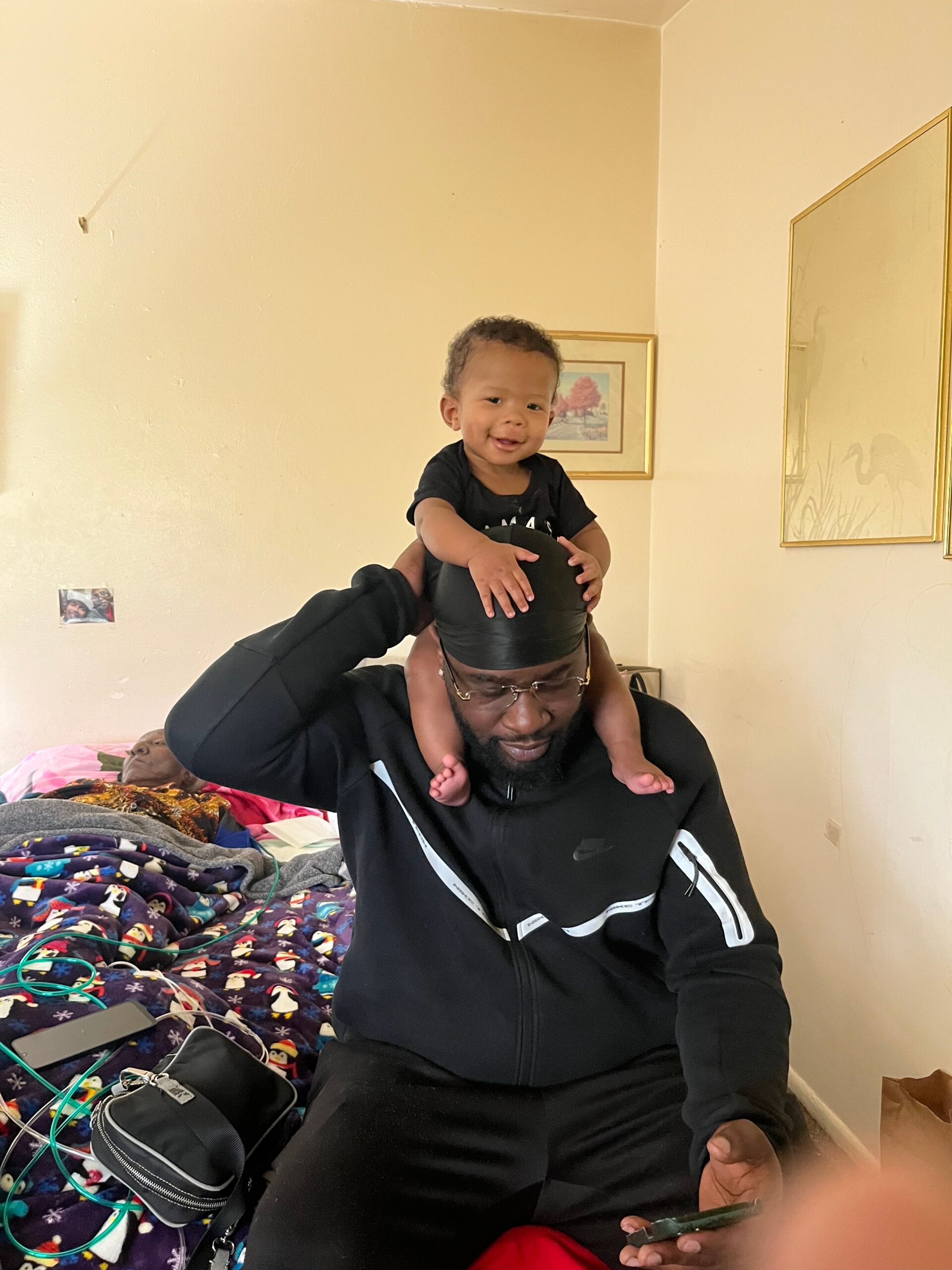 A man with a baby on his shoulders sits in a messy bedroom filled with blankets, cables, and personal items on a bed. The child smiles at the camera.