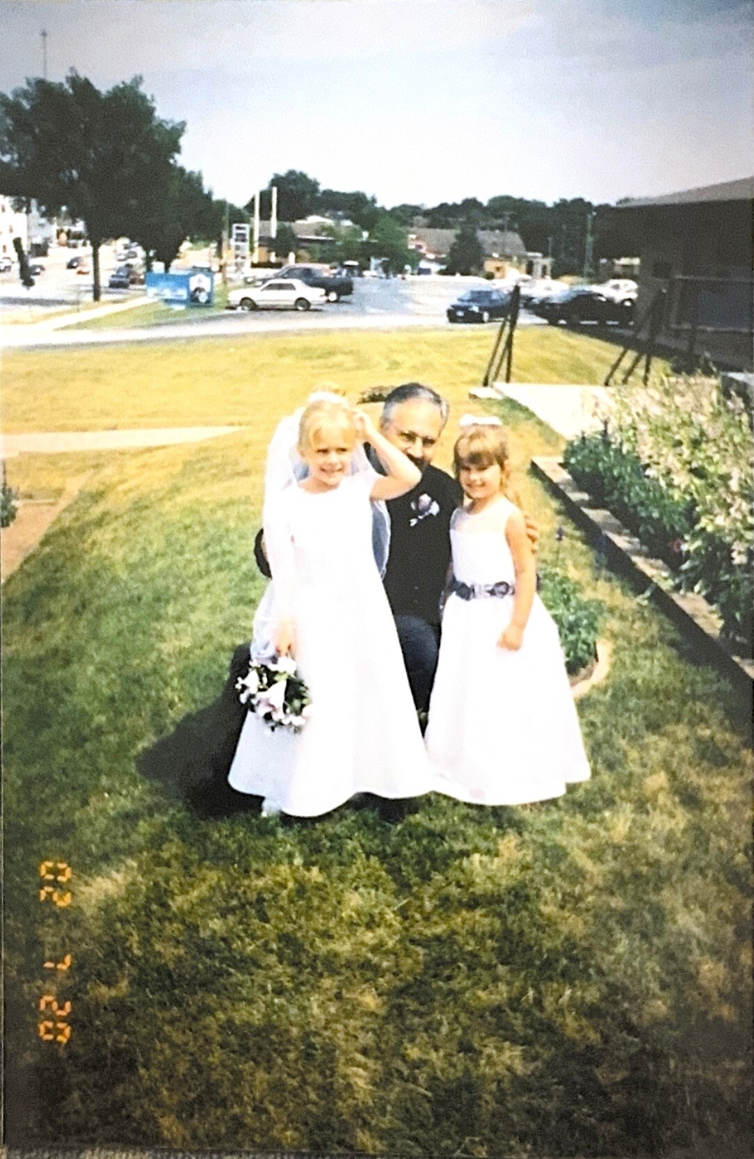 Man kneels on a grassy lawn with two young girls in white dresses, one holding a bouquet, with a street scene in the background.