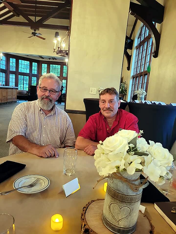 Two middle-aged men sit at a restaurant table with a white flower centerpiece and small candles on the table in a warmly lit room.