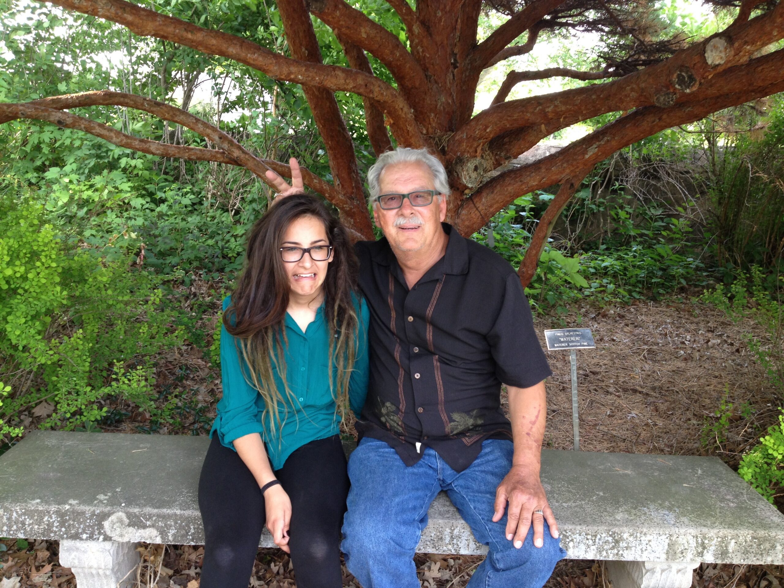 Young woman and older man posing on a bench outdoors under a large tree; she’s making bunny ears behind his head.