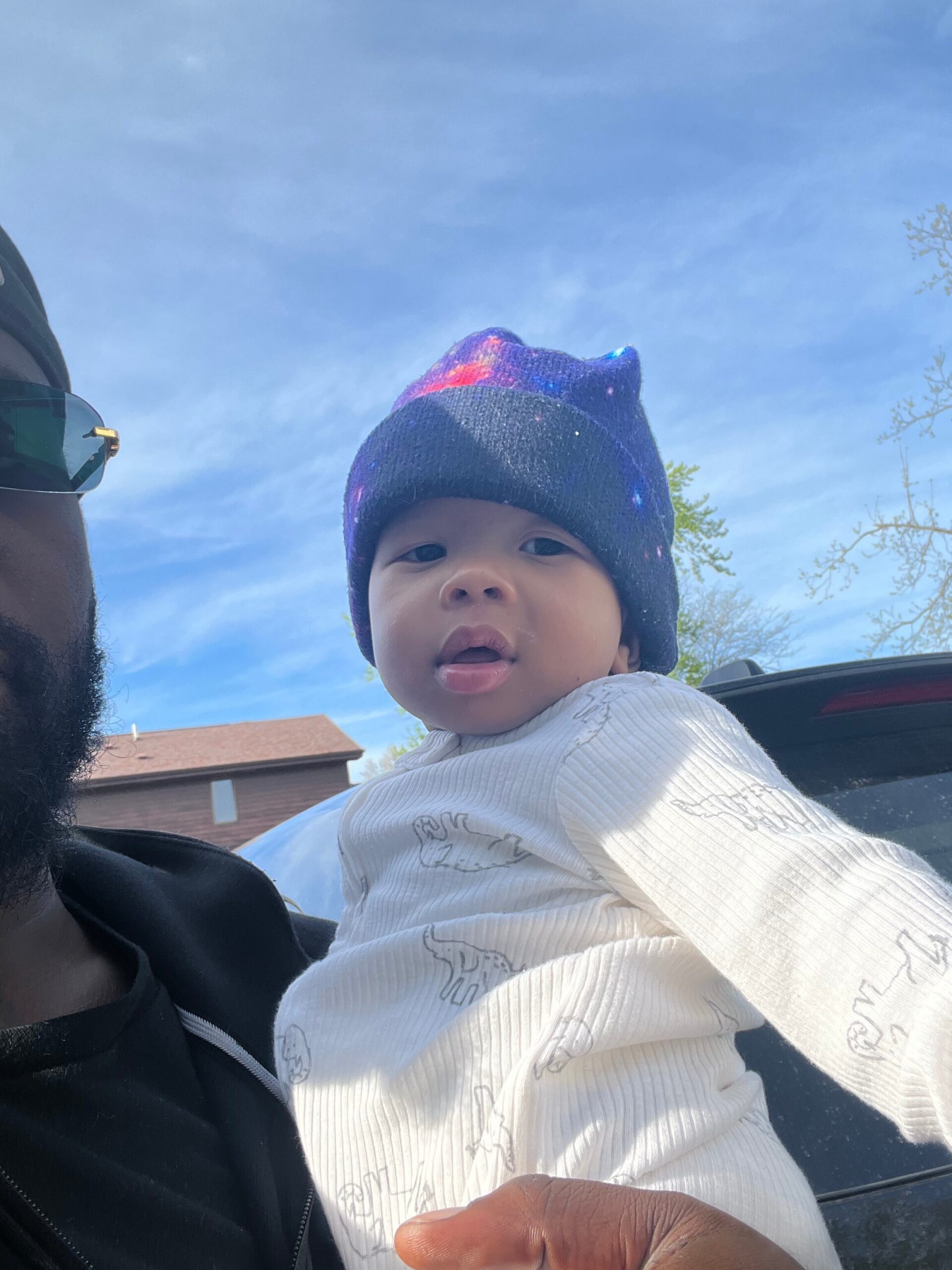 Baby being held outdoors, wearing a navy knit hat with colorful specks, against a clear blue sky; part of an adult with sunglasses is visible on the left.