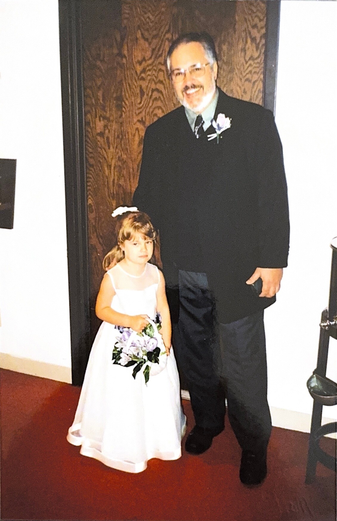 Smiling man in a dark suit with a boutonniere standing beside a young girl in a white dress holding a bouquet at a formal event.