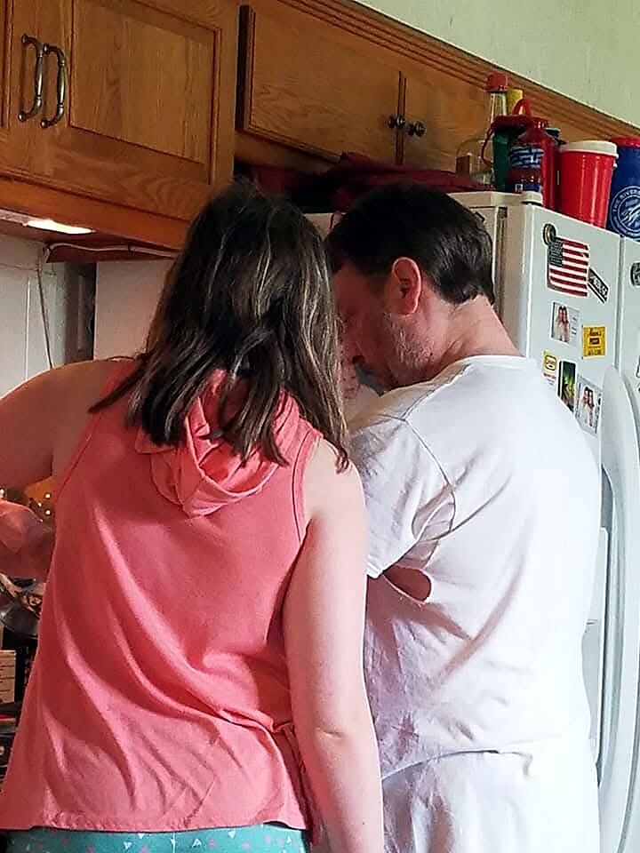 Two people seen from behind standing at a kitchen counter near a refrigerator, preparing food together.