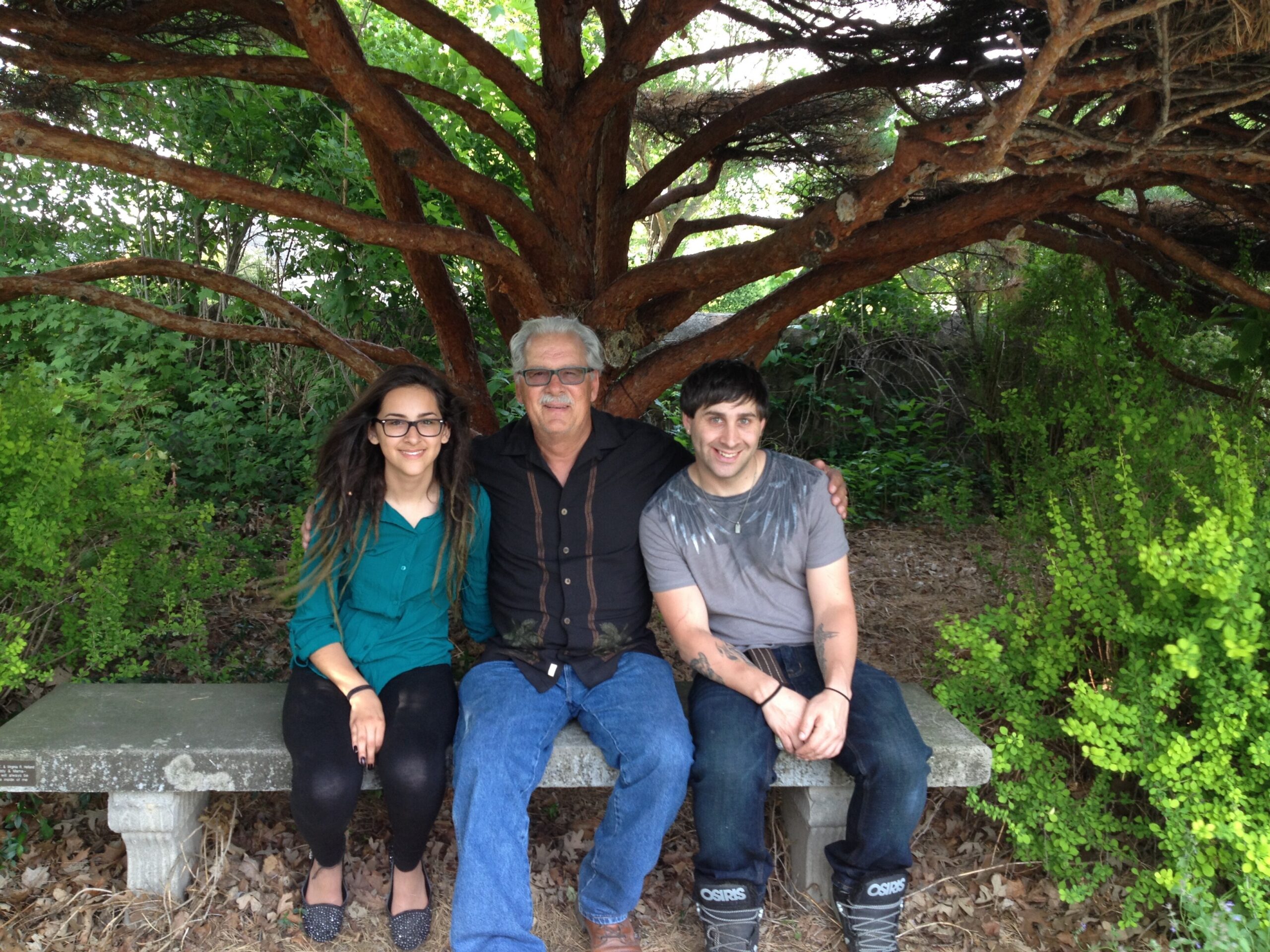 Three people sit on a stone bench outdoors under a large tree, smiling at the camera.] ,