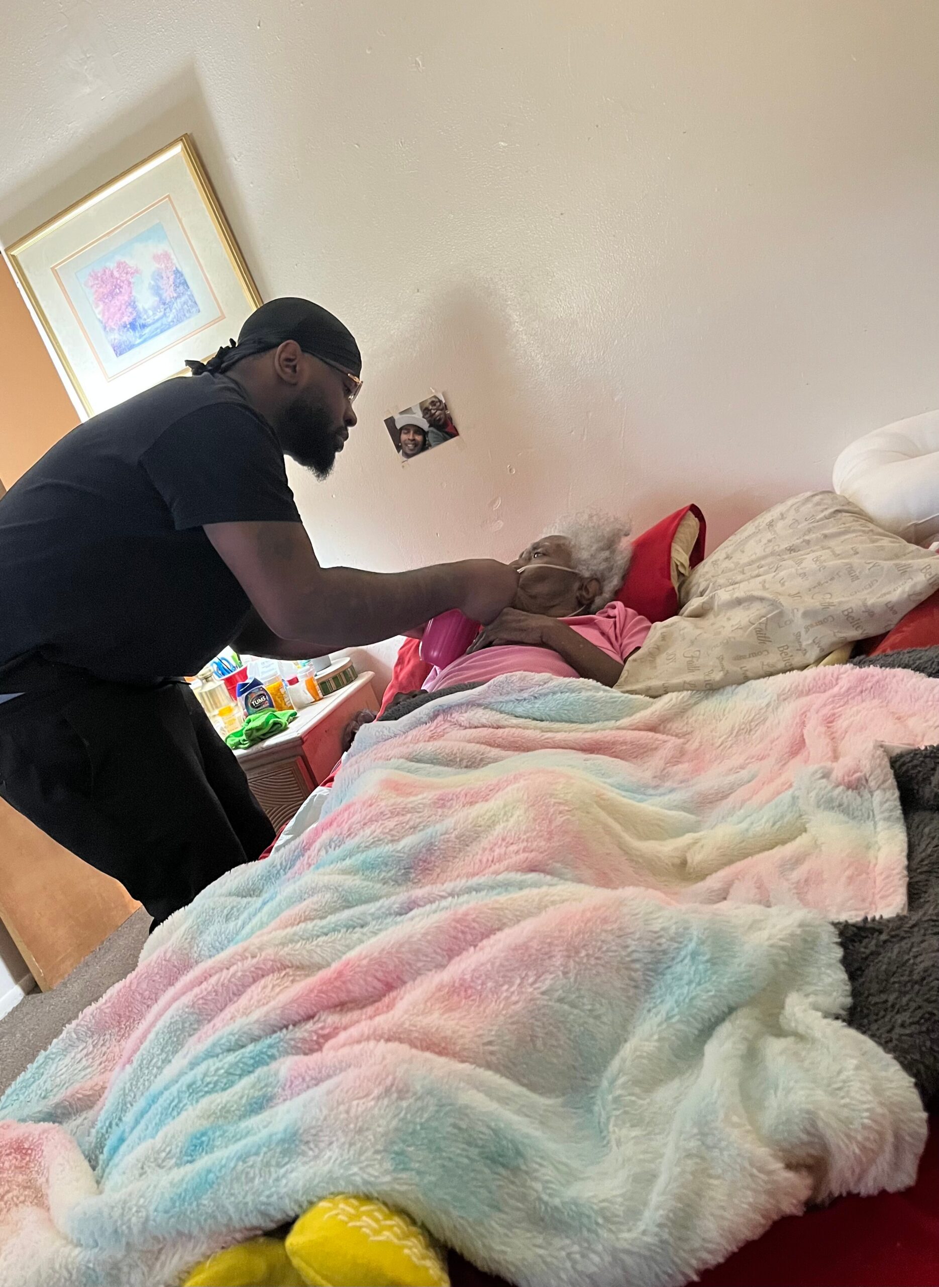 Caregiver in a dark shirt and durag leaning over an elderly person in bed, holding their hands under pastel blankets at a home bed setup.