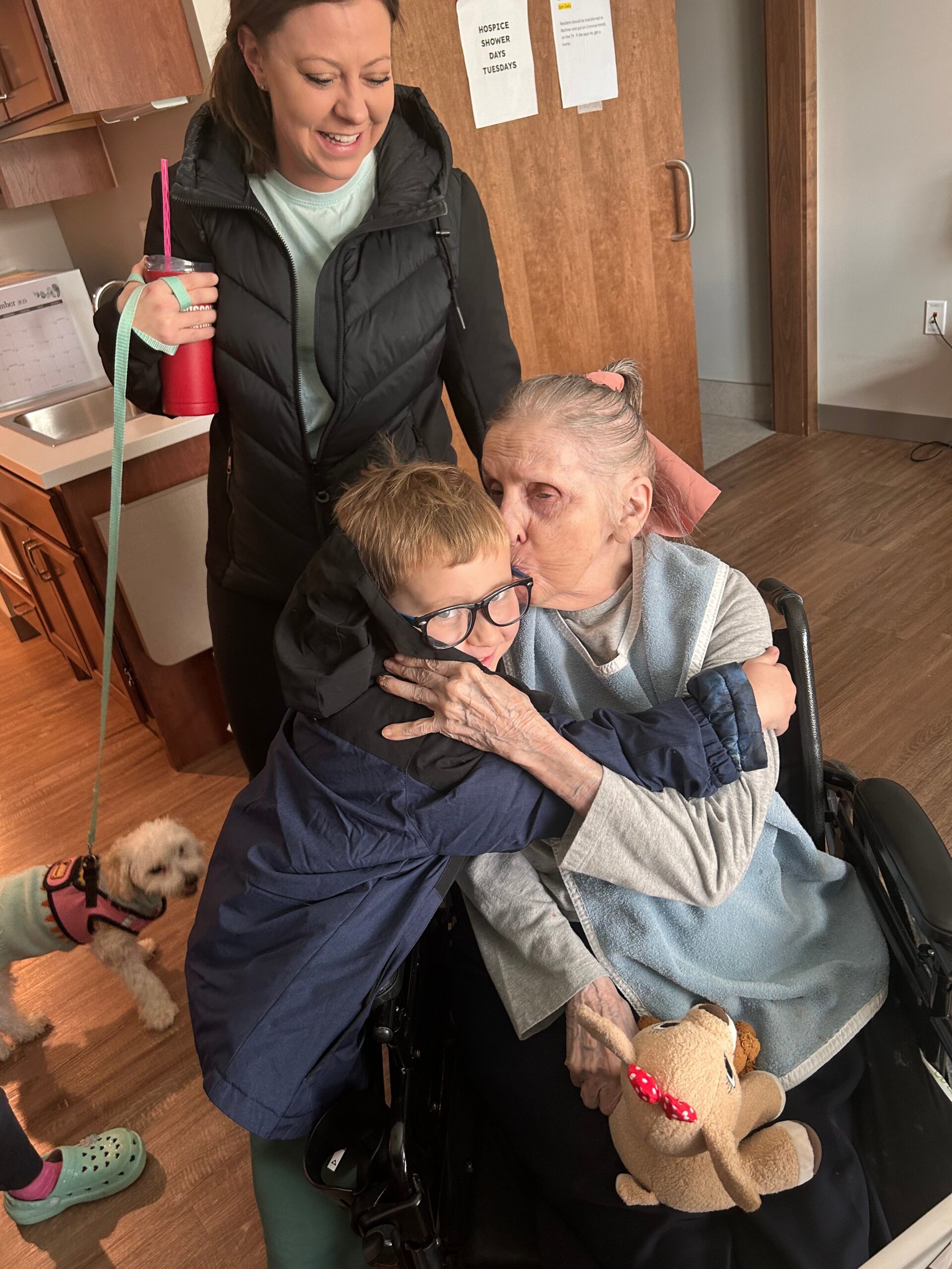 Caregiver smiles while standing beside an elderly woman in a wheelchair, who hugs a young boy; a small dog in a sweater sits on the floor nearby.