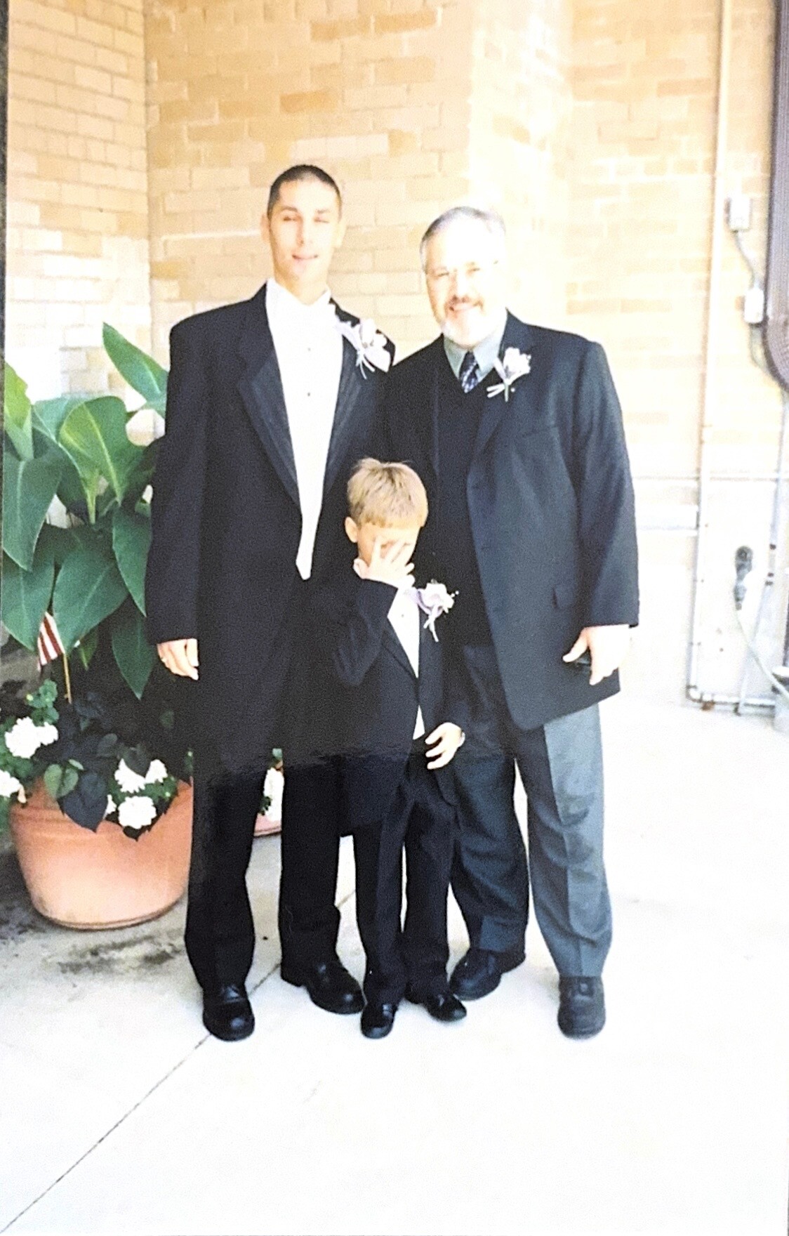 Three men in black tuxedos posing outside a brick wall, with a boy in front covering his face.