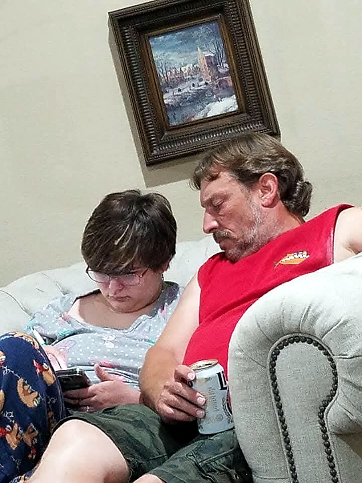 Man in a red sleeveless shirt sits on a sofa next to a woman looking at her phone; a framed painting hangs on the wall behind them.